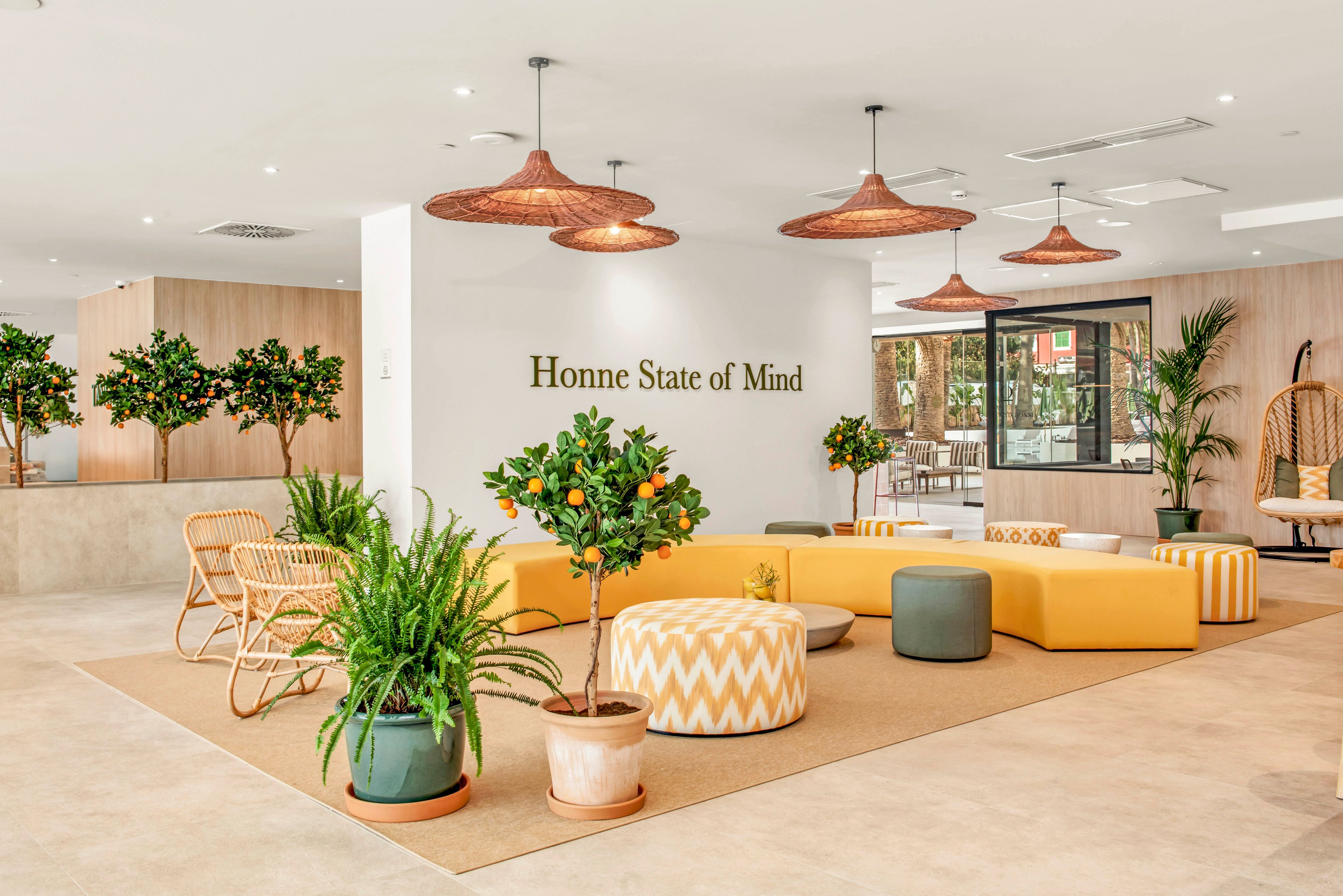 Contemporary lounge area featuring yellow modular seating and patterned ottomans is set within a spacious, light-filled interior. Potted citrus trees and lush green plants add natural color and freshness throughout the room. Woven rattan chairs, hanging light fixtures, and neutral flooring create a relaxed, resort-like atmosphere. The wall text reads "Honne State of Mind," reinforcing a calm, mindful mood.