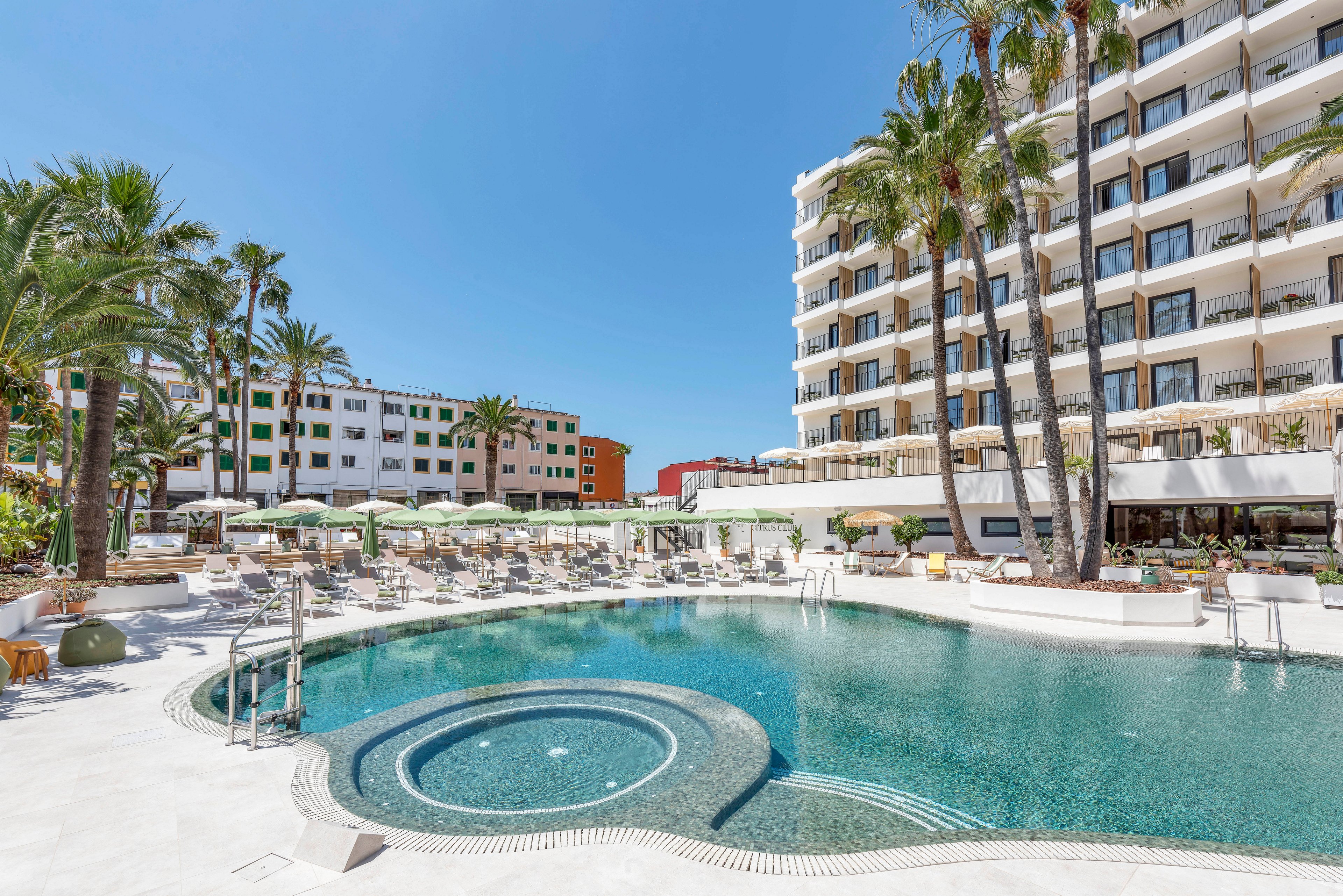 Outdoor view of a resort-style swimming pool in front of a multi-story hotel building. The scene features curved pool edges, a circular whirlpool area, and metal handrails leading into the water. Numerous sun loungers and umbrellas line the pool deck, surrounded by tall palm trees and other greenery. Clear blue sky and bright sunlight create a relaxed vacation atmosphere in an urban coastal setting.