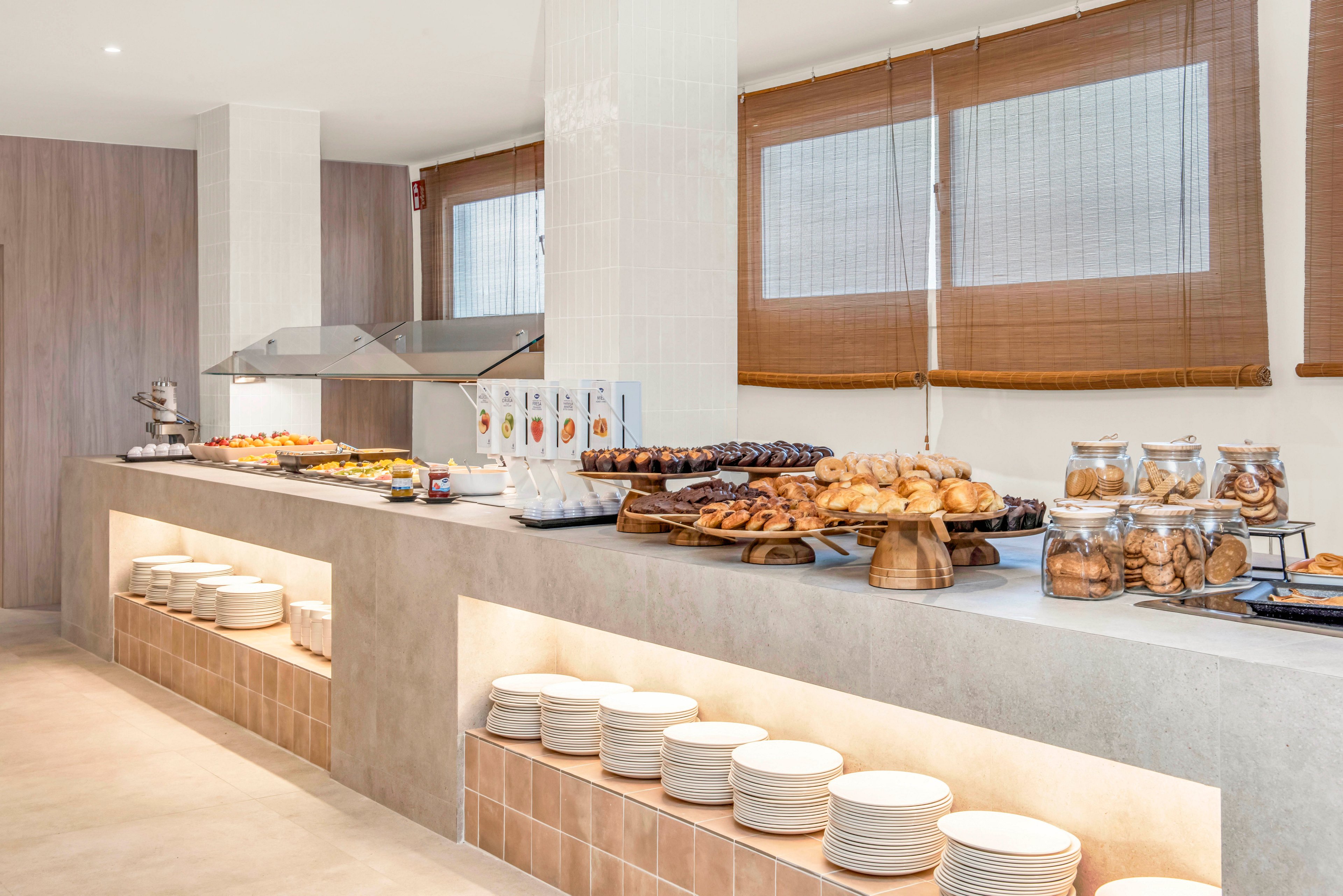 A bright, modern buffet counter displays a wide variety of pastries and baked goods. The setting appears to be a hotel or restaurant breakfast area with clean, minimalist decor. Stacks of white plates are neatly arranged on open shelves beneath the counter, ready for guests. Glass jars and covered trays showcase cookies, croissants, and other treats, while warm neutral tones and natural light create an inviting atmosphere.