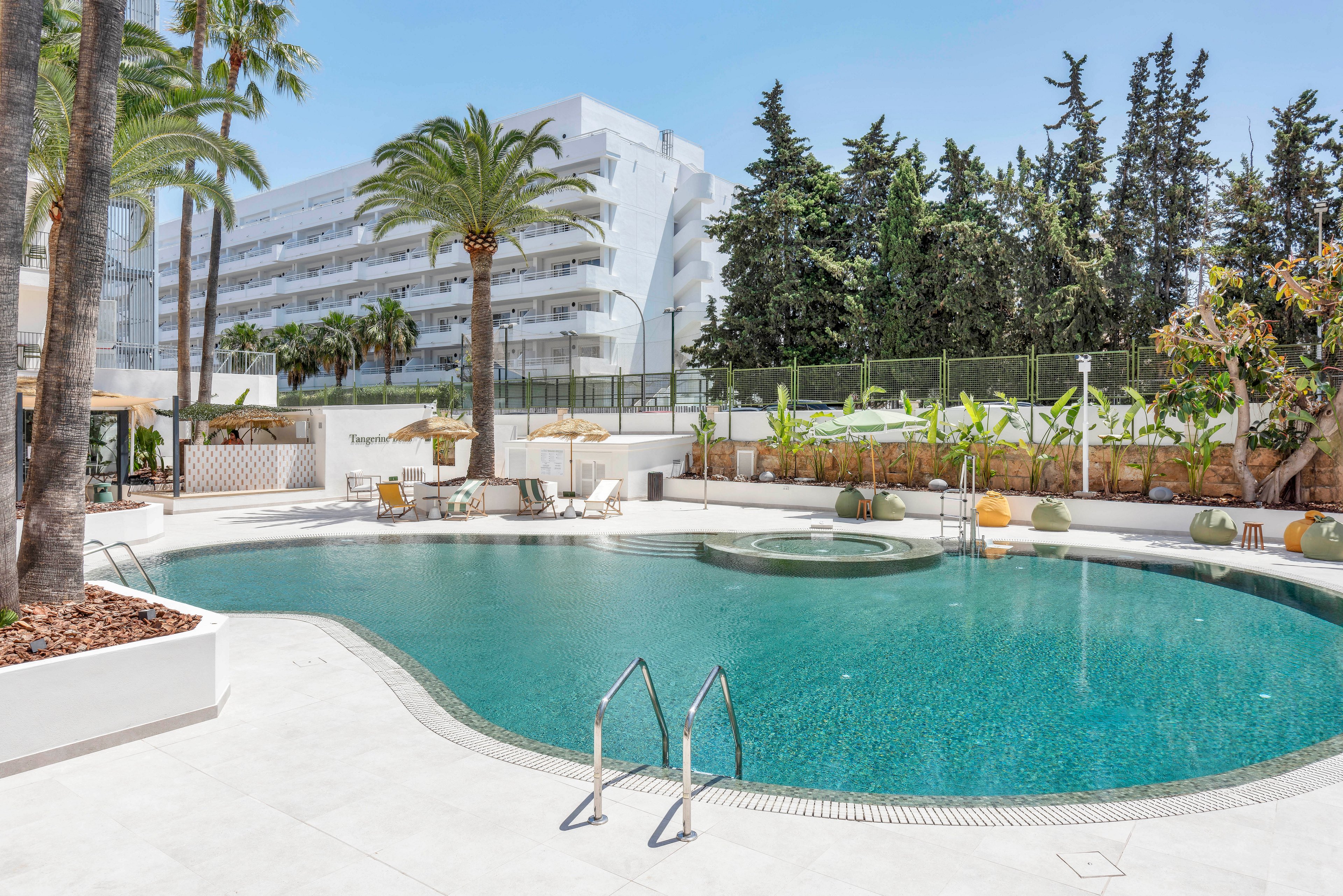A curved outdoor swimming pool sits in front of a modern white hotel building. Palm trees and lush greenery surround the pool area, creating a bright and relaxing vacation atmosphere. Sun loungers and umbrellas are arranged around the deck, suggesting a leisure and resort setting. The scene is captured on a clear day with soft reflections on the turquoise water.