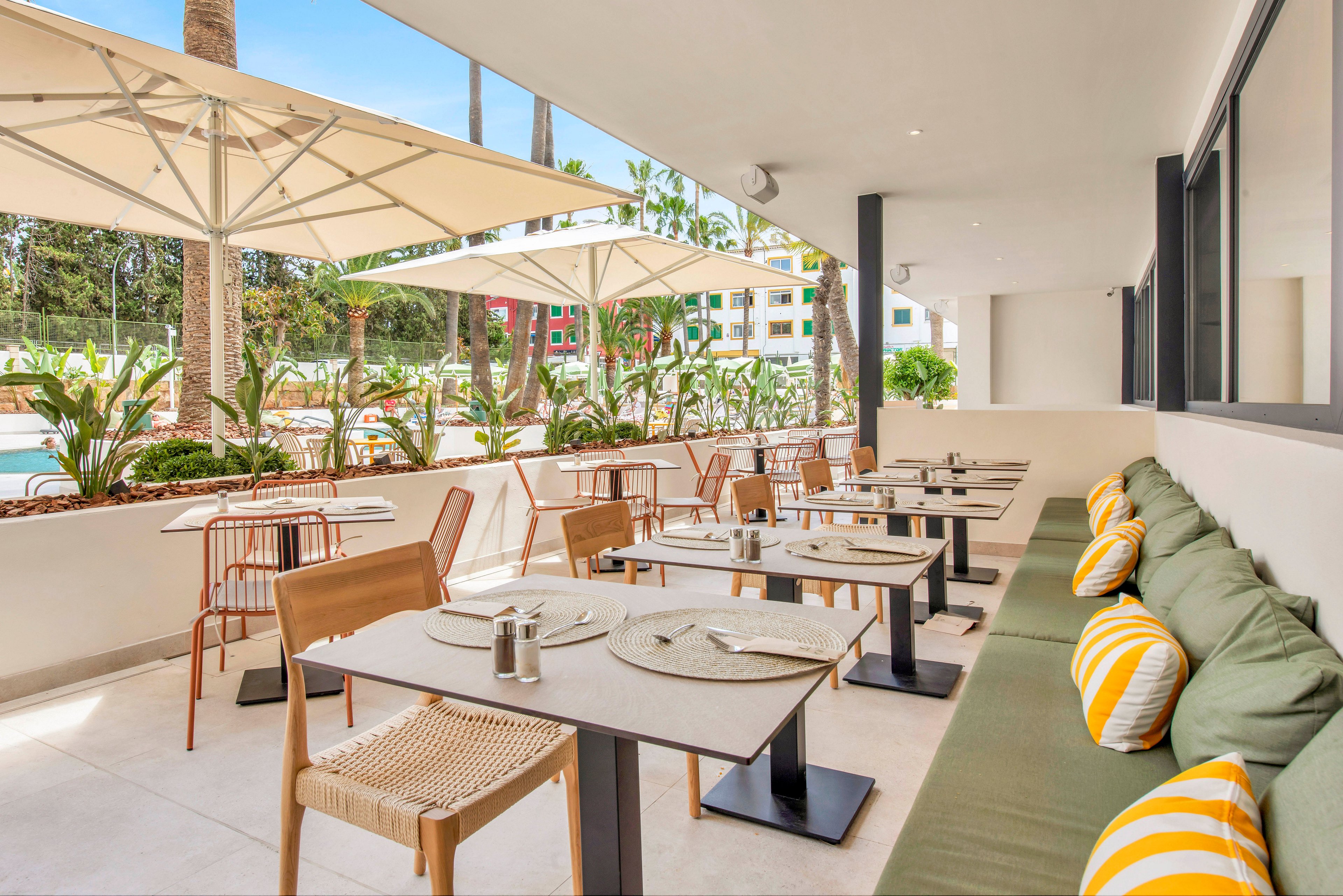 Bright outdoor restaurant terrace with neatly arranged tables and chairs. A long green cushioned bench with yellow and white striped pillows lines one side, facing modern dining tables set with placemats and condiments. Large umbrellas provide shade, while tropical plants and palm trees frame the open-air space. In the background, a pool area and colorful buildings suggest a relaxed vacation setting.