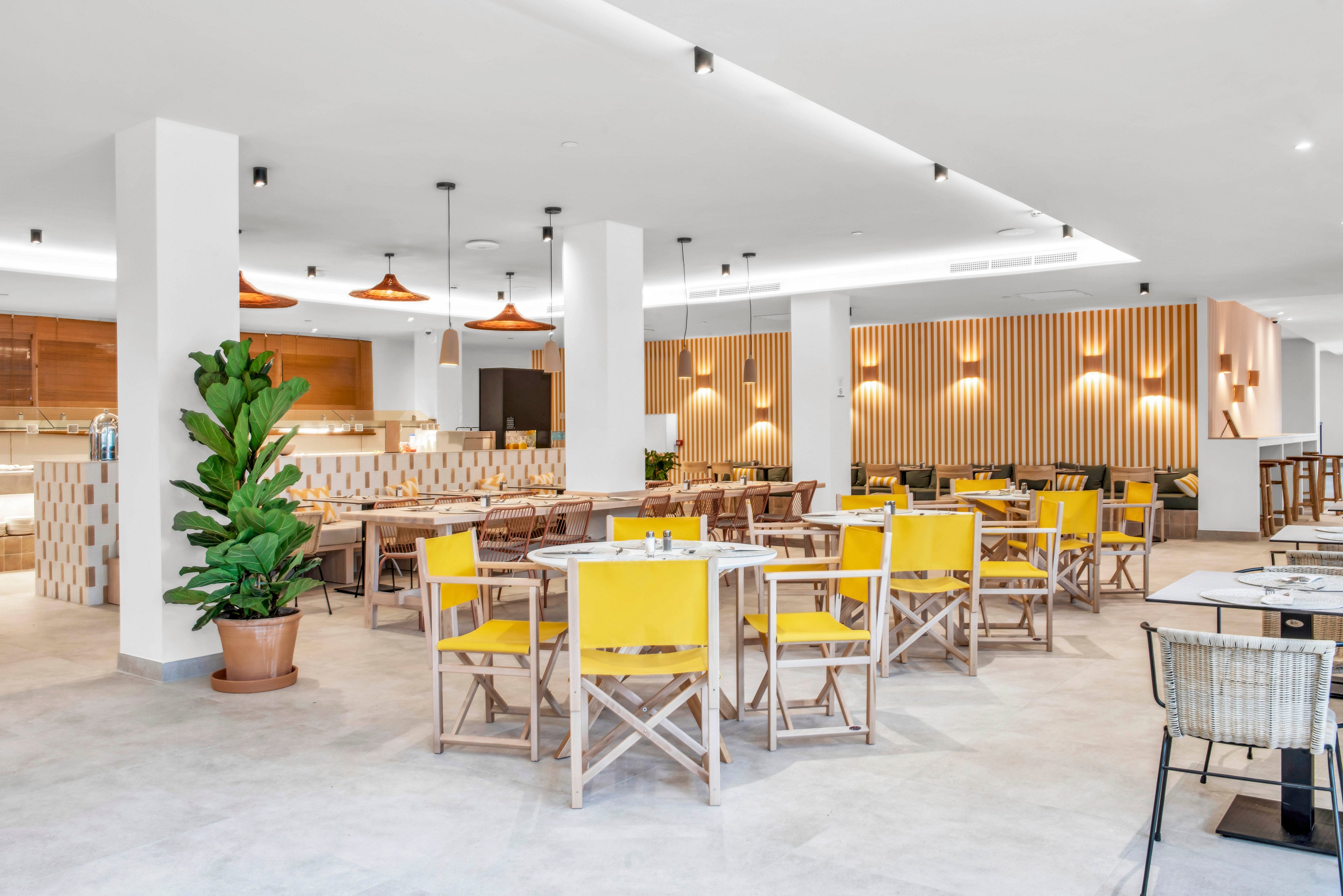 Contemporary restaurant dining area featuring wooden tables and yellow chairs is shown in a spacious, open layout. Warm pendant lights hang from the ceiling, illuminating the bar and service counter in the background. Light neutral flooring and vertical wooden slats on the wall create a clean, minimalist aesthetic. A large potted plant adds a touch of greenery to the bright, inviting space.