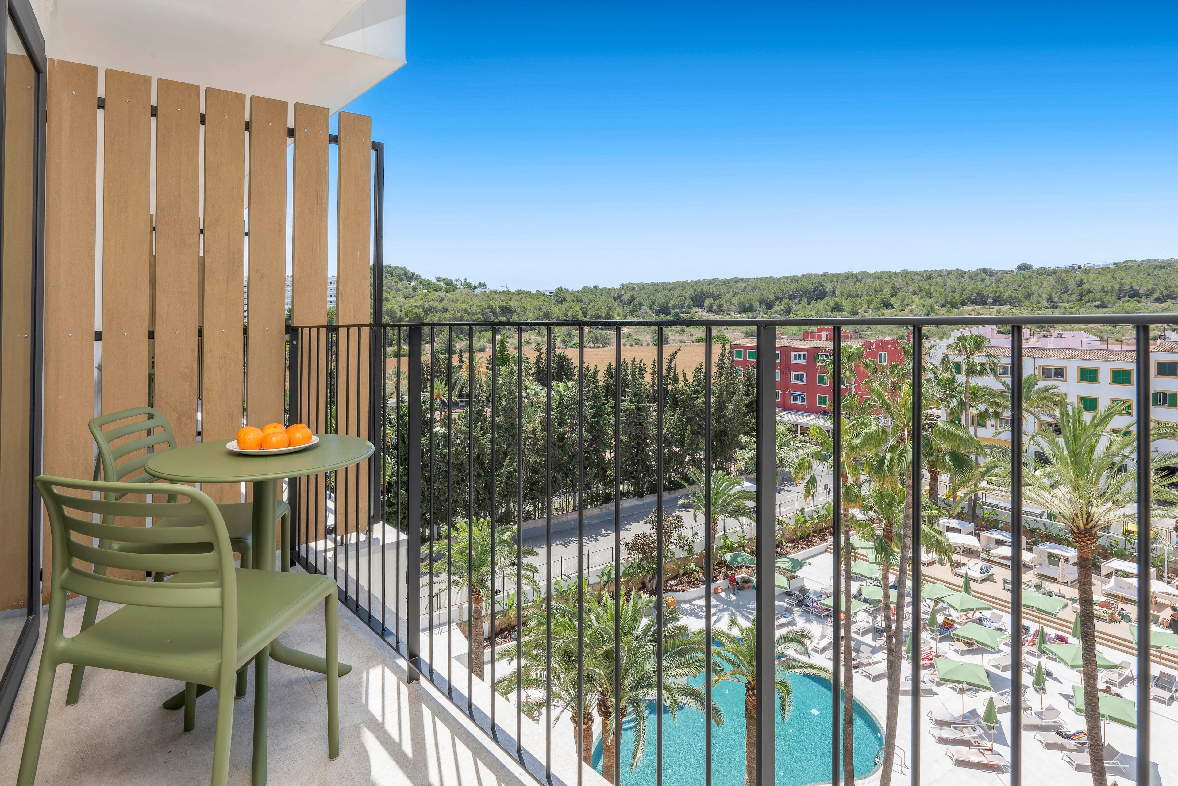 A modern hotel balcony features a small green table with matching chairs and a bowl of oranges. Black railings frame the view of a resort-style pool area below with sun loungers and palm trees. Beyond the pool, colorful buildings stand against a backdrop of dense green forest. The bright blue sky and strong sunlight create a fresh, inviting vacation atmosphere.