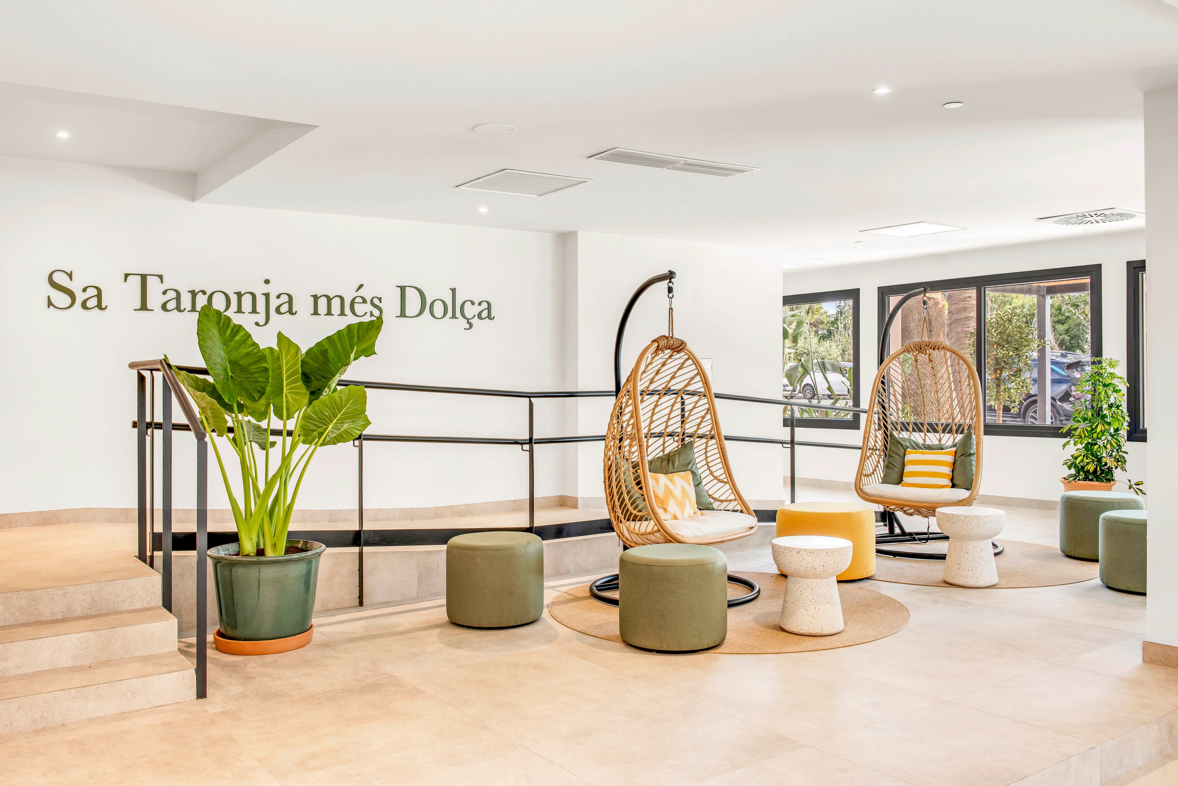 Bright contemporary lounge area featuring two hanging wicker chairs with cushions. The space includes low poufs, small side tables, and large potted green plants on a light tiled floor. A black metal railing separates the seating zone from the steps, and large windows bring in natural light and outdoor views. Text on the wall reads “Sa Taronja més Dolça,” adding a decorative focal point in the room.
