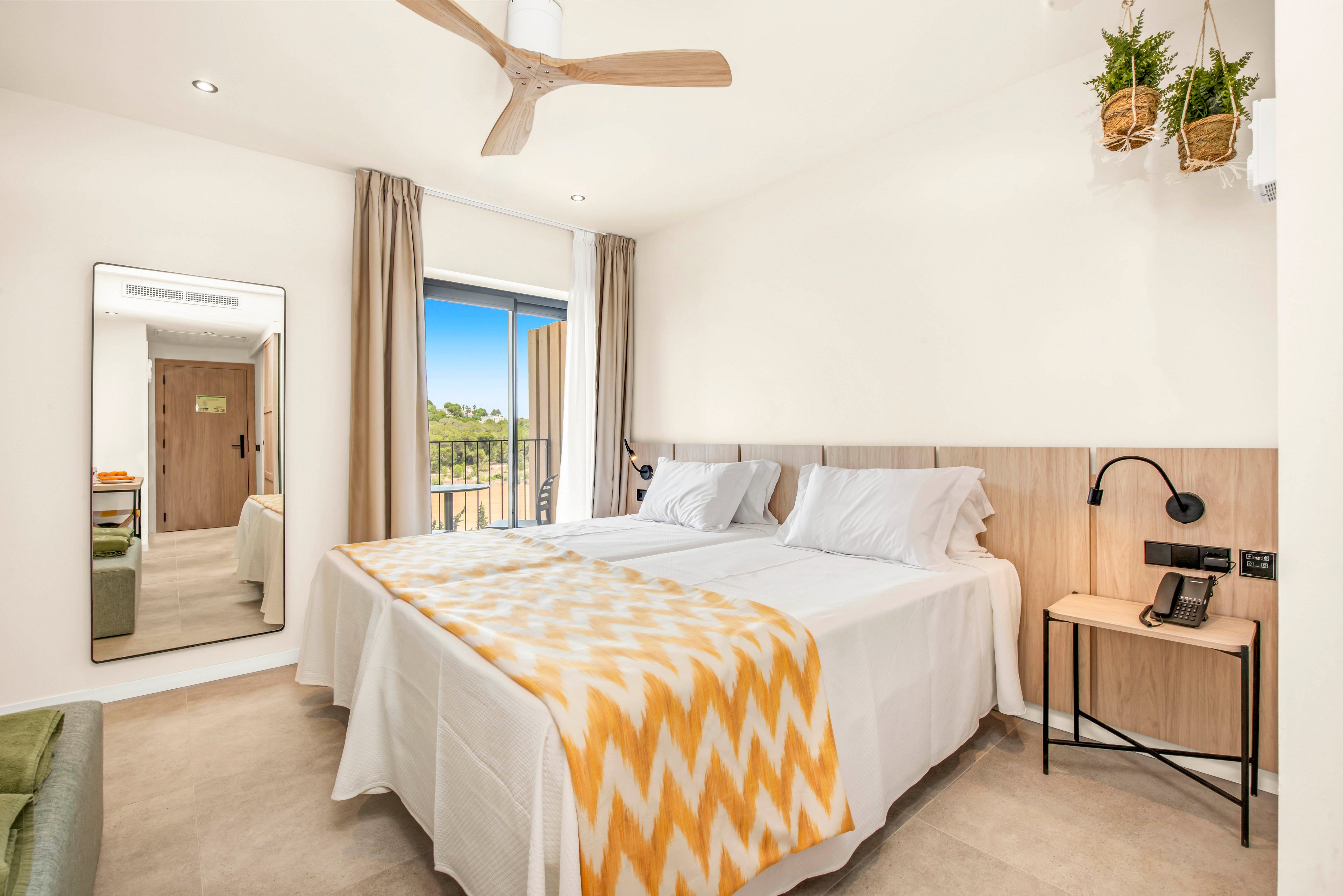 A contemporary double bedroom features two single beds pushed together with white linens and a yellow chevron blanket. The room has light wood accents, a bedside table with a telephone, and wall-mounted reading lights. A large mirror reflects the entrance area, while a ceiling fan and hanging plants add comfort and decor. Sunlight streams in through the balcony door, revealing an outdoor view of greenery and blue sky.