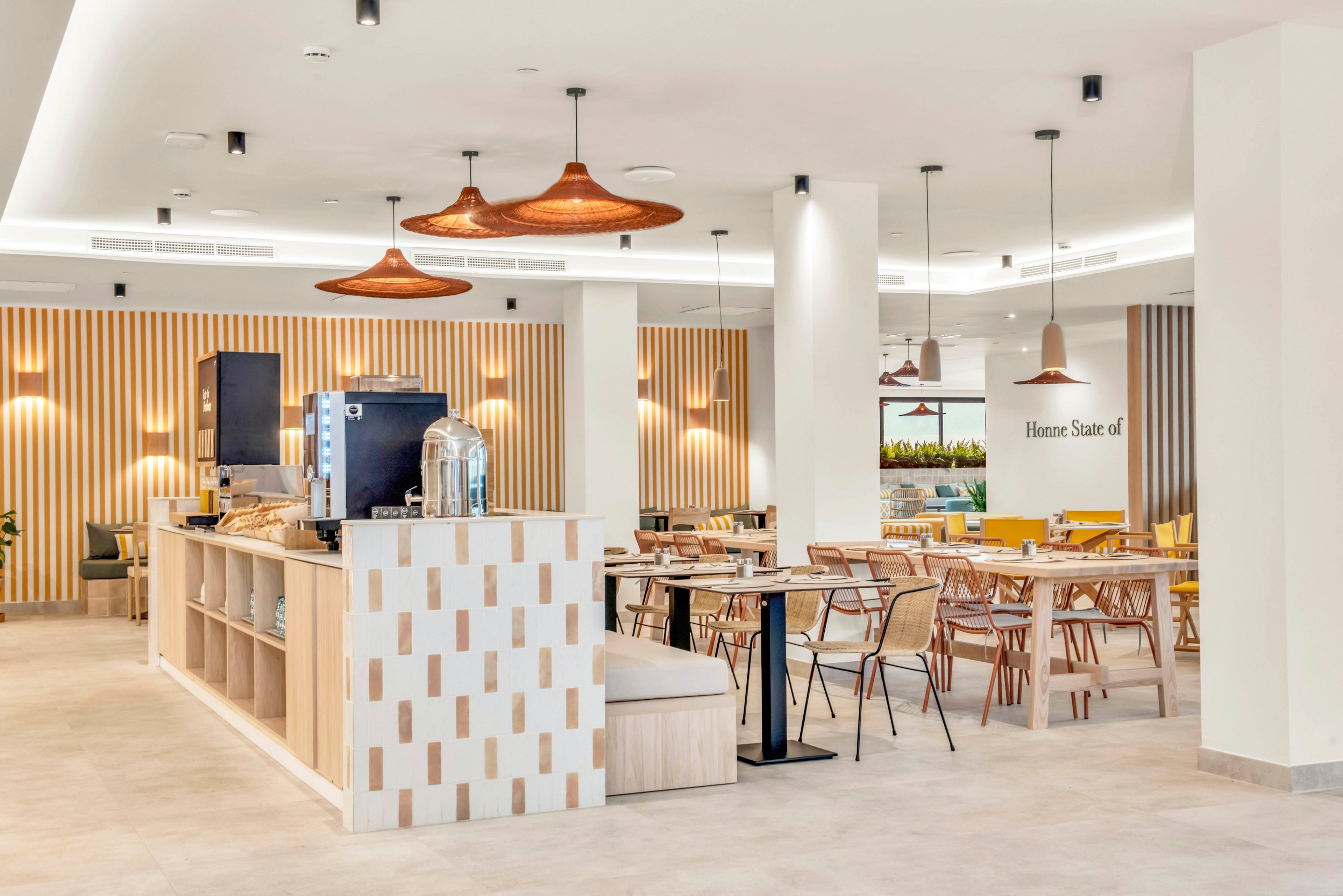 Contemporary café interior featuring wooden tables and mixed seating in a spacious, bright room. A central counter with beverage dispensers and equipment stands in the foreground. Warm pendant lights and vertical striped wall panels add texture and visual interest. The space feels clean, inviting, and minimalist, with neutral tones accented by soft yellows and natural wood.