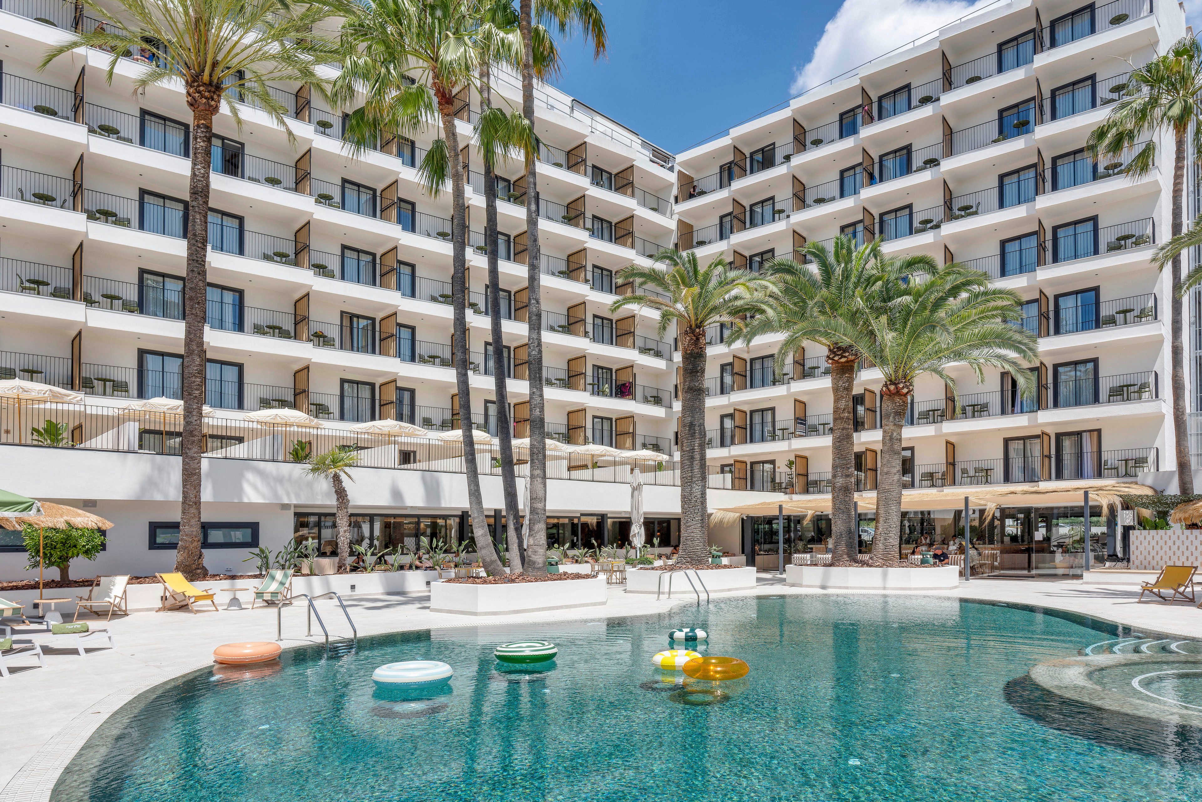 A contemporary multi-story resort hotel overlooks a circular outdoor swimming pool. Palm trees and sun loungers surround the pool, creating a relaxed vacation atmosphere. Colorful inflatable pool floats rest on the water, while balconies and terraces are visible across the building facade. Bright daylight and clear skies enhance the sunny, Mediterranean-style setting.