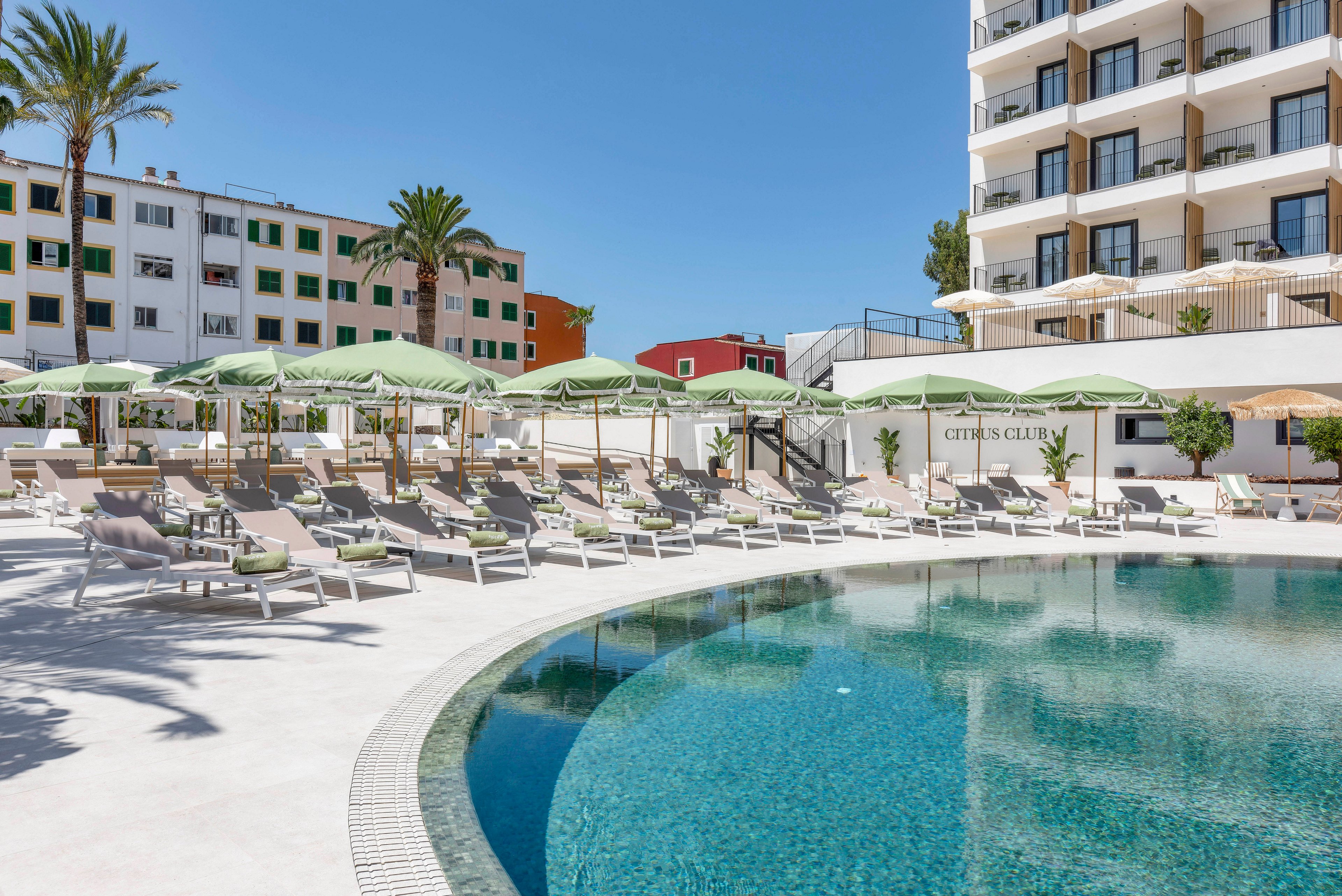 A curved outdoor swimming pool is surrounded by rows of sun loungers and green umbrellas at a modern resort. Multi-story buildings with balconies rise in the background under a clear blue sky. Palm trees and neatly arranged seating create a relaxed vacation atmosphere. The scene suggests a warm, sunny day ideal for leisure and sunbathing.