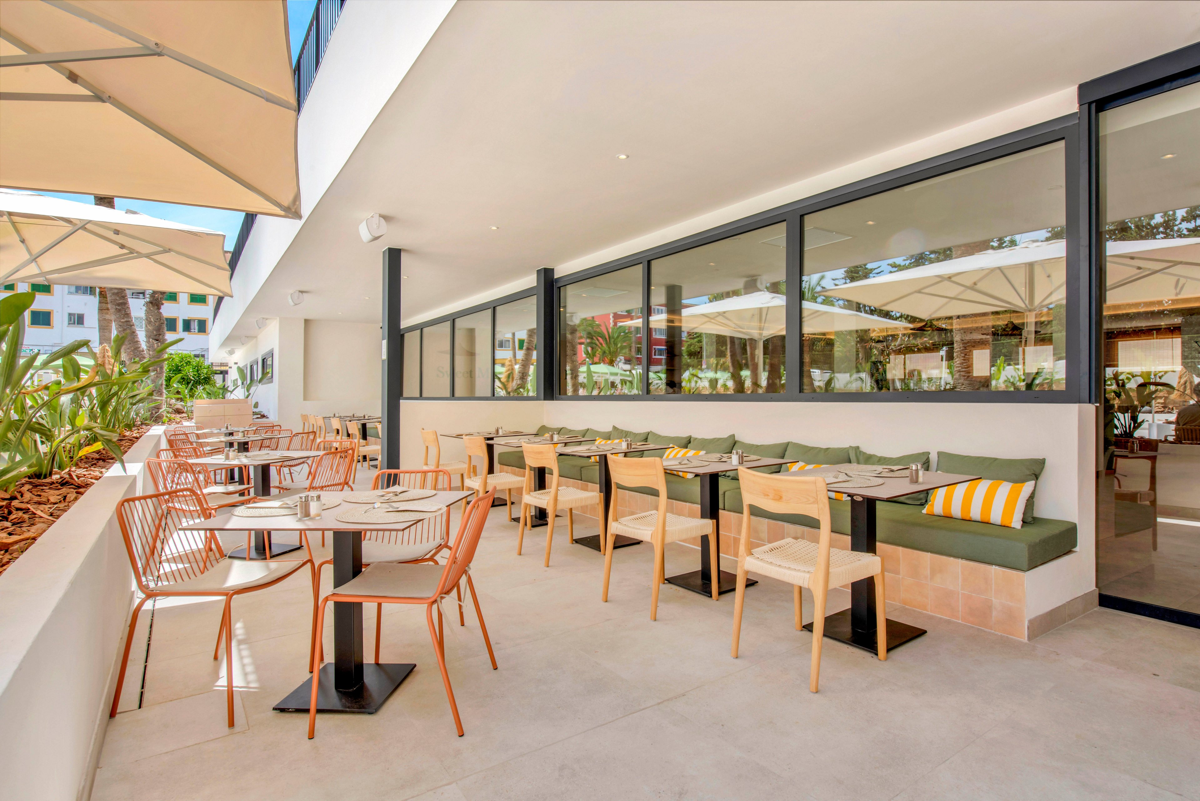 Contemporary restaurant terrace with neatly arranged tables and chairs is shown in bright daylight. The setting features an open-air patio beside large glass windows, with cushioned bench seating and striped throw pillows. Neutral tones, clean lines, and potted plants create a relaxed, modern atmosphere. Sun umbrellas provide shade, suggesting a comfortable space for outdoor dining and socializing.