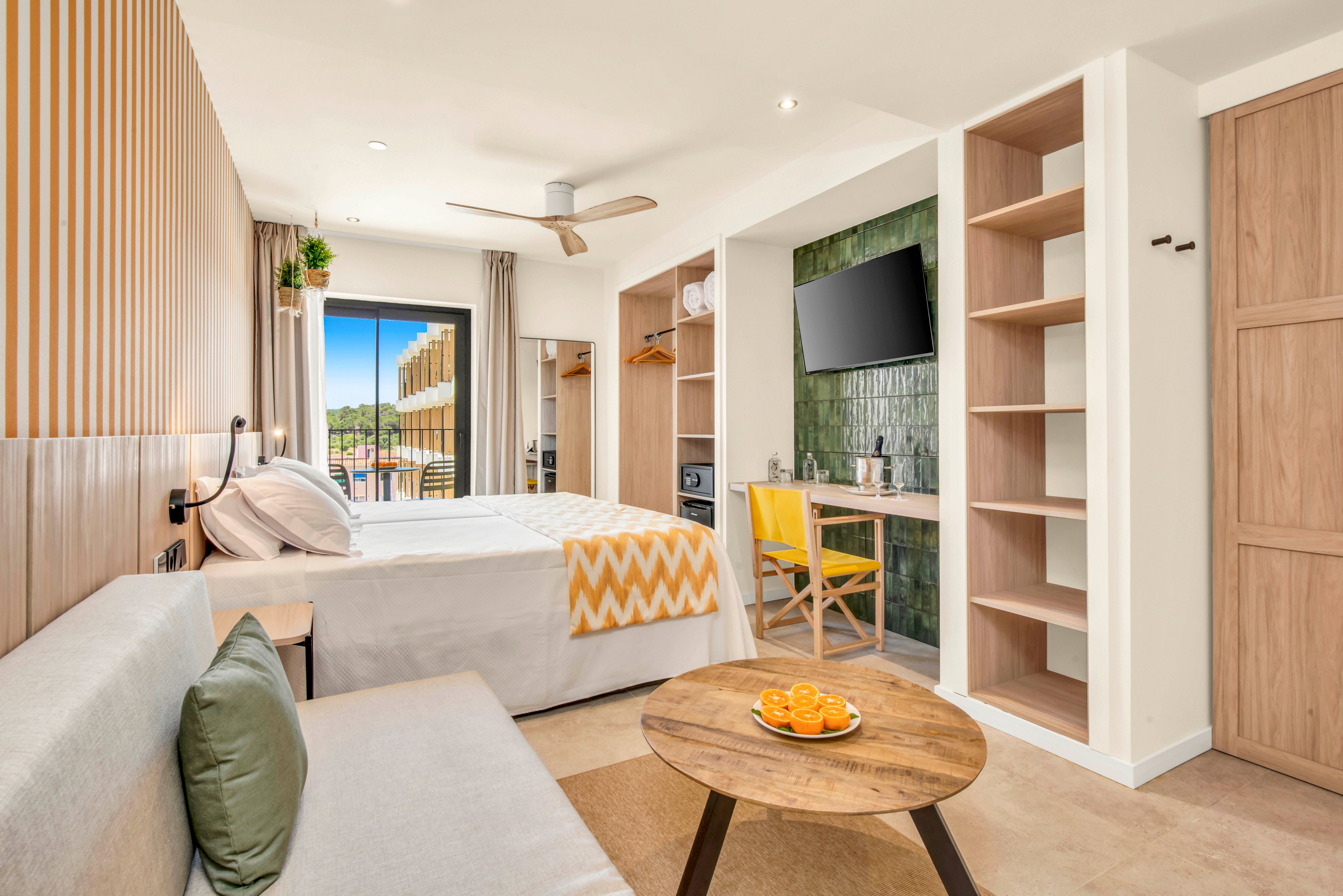 Contemporary hotel room interior featuring a neatly made double bed with a yellow chevron throw. A sofa and round wooden coffee table with sliced oranges create a cozy seating area. The space includes open shelving, a wall-mounted television, and a small desk with a yellow chair. Natural light streams in through the balcony door, revealing an outdoor view with trees and buildings.