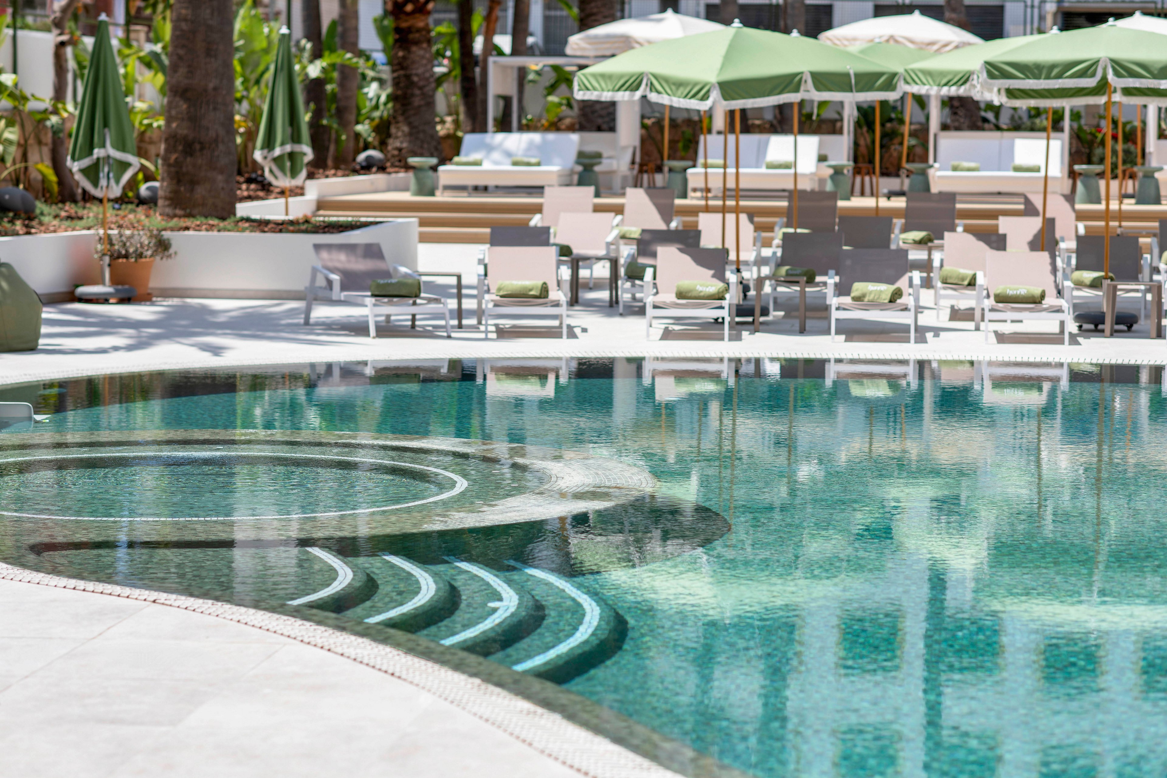 A clear turquoise swimming pool with curved steps sits in the foreground of a modern outdoor resort area. Sun loungers with cushions and side tables are arranged around the pool under green and white umbrellas. Palm trees and lush plants add a tropical feel, while white buildings and terraces frame the scene. The bright daylight and soft colors create a relaxed vacation atmosphere.
