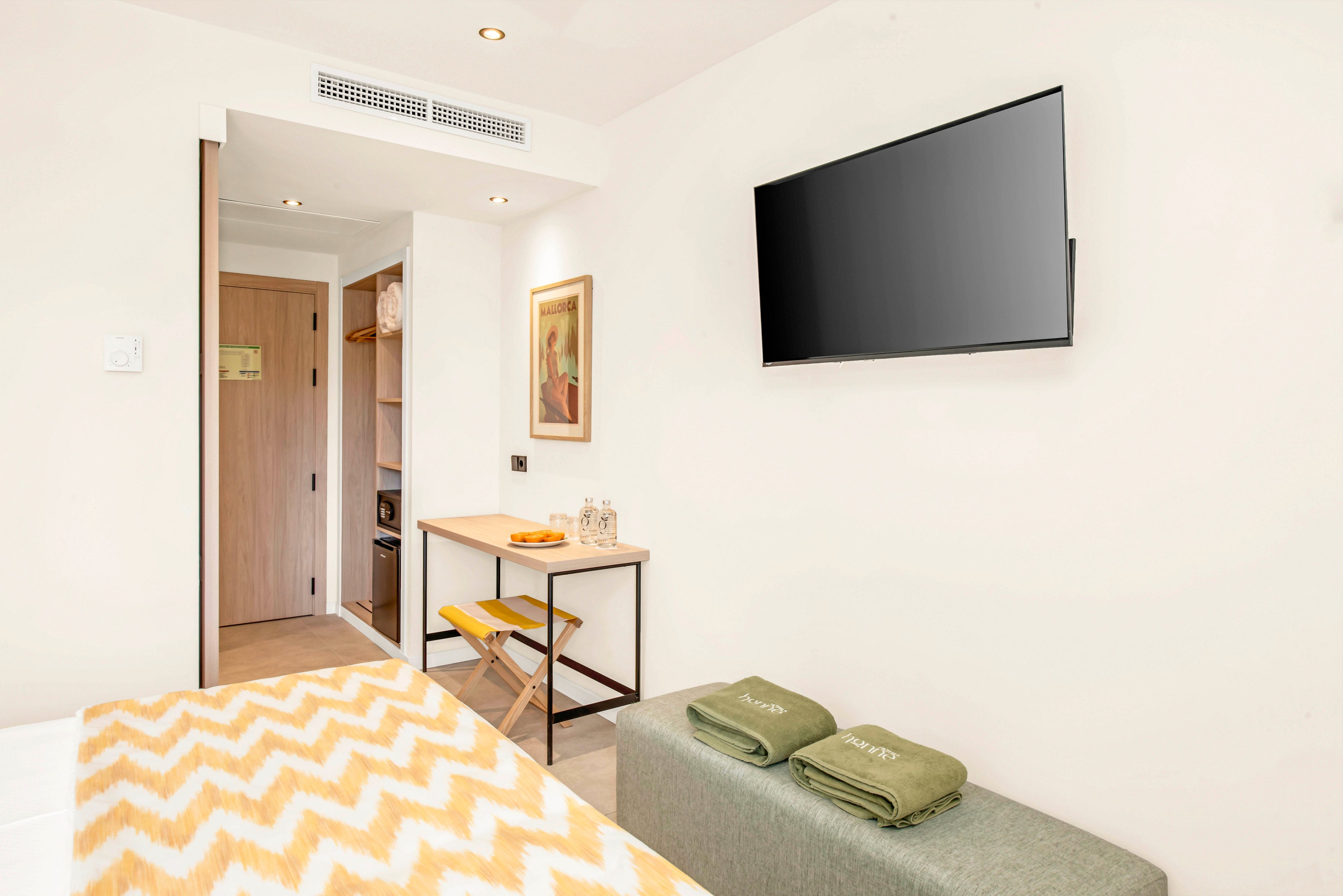 Bright modern hotel room interior featuring a wall-mounted flat-screen television. A small wooden desk with a chair and decorative items sits near the doorway and built-in shelving. In the foreground, a bed with a yellow and white chevron blanket and a cushioned bench with neatly folded green towels add color and comfort. The overall space appears clean, minimal, and contemporary with light walls and wood accents.