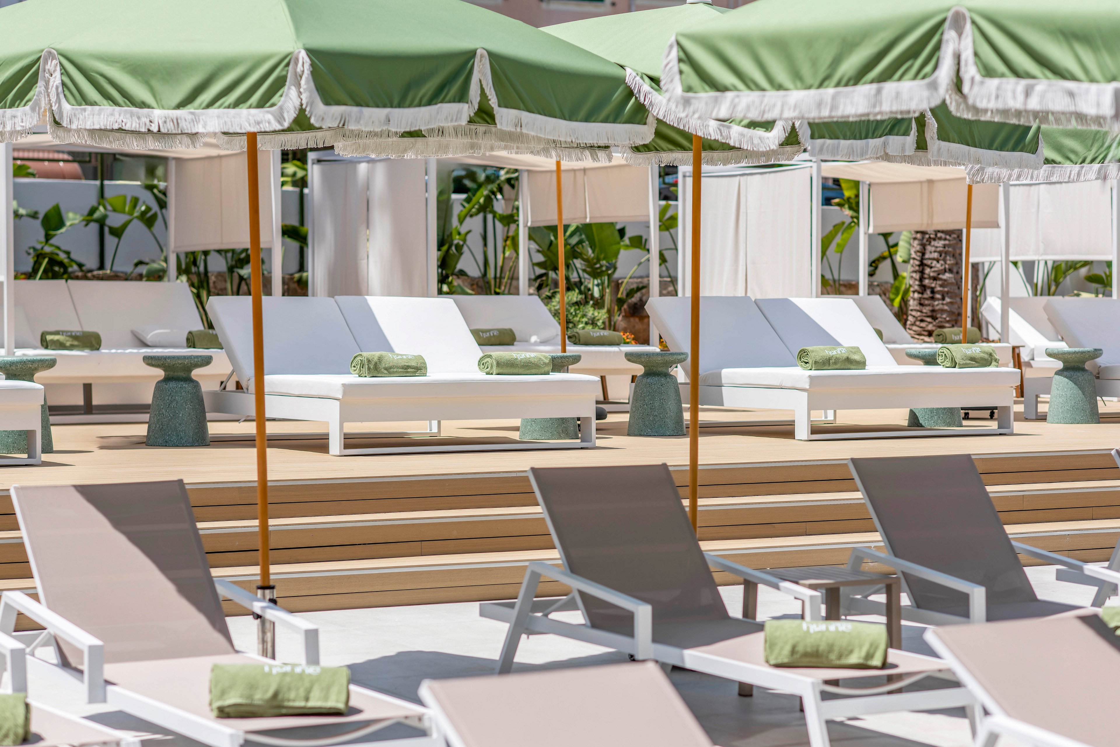 The image shows a stylish outdoor poolside area with white sun loungers arranged in neat rows. Behind them, elevated daybeds with green cushions sit under large green-and-white umbrellas, creating a resort-like atmosphere. Light wood decking, small side tables, and tropical plants in the background add to the relaxed, vacation setting. The overall scene feels bright, clean, and inviting, ideal for leisure and sunbathing.