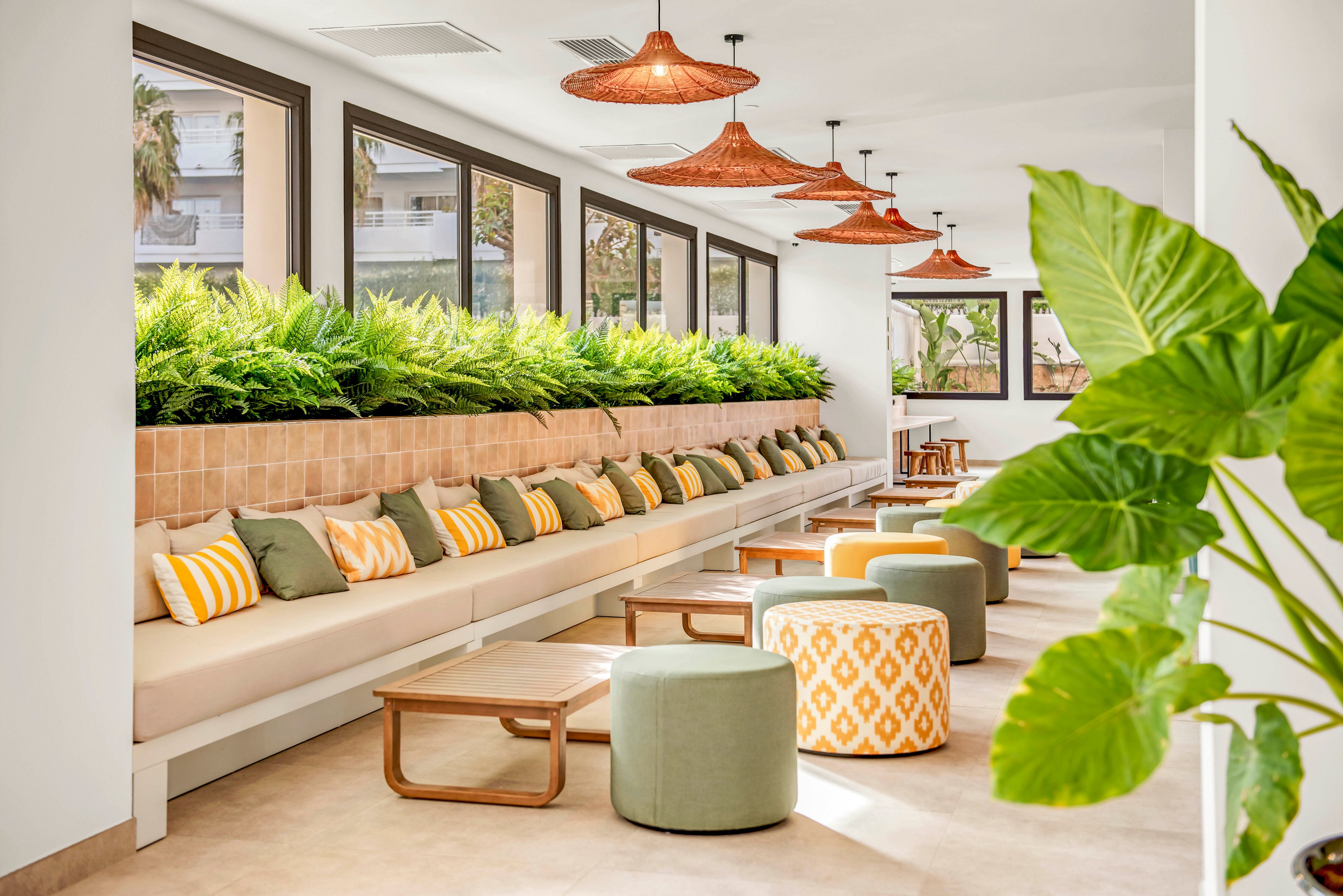 Contemporary indoor lounge area featuring a long built-in bench lined with colorful cushions. Green plants in a raised planter add a lush natural accent along the wall of large windows. Round upholstered poufs and low wooden tables create a casual seating arrangement. Warm neutral tones with green and yellow accents give the space a relaxed, inviting atmosphere.
