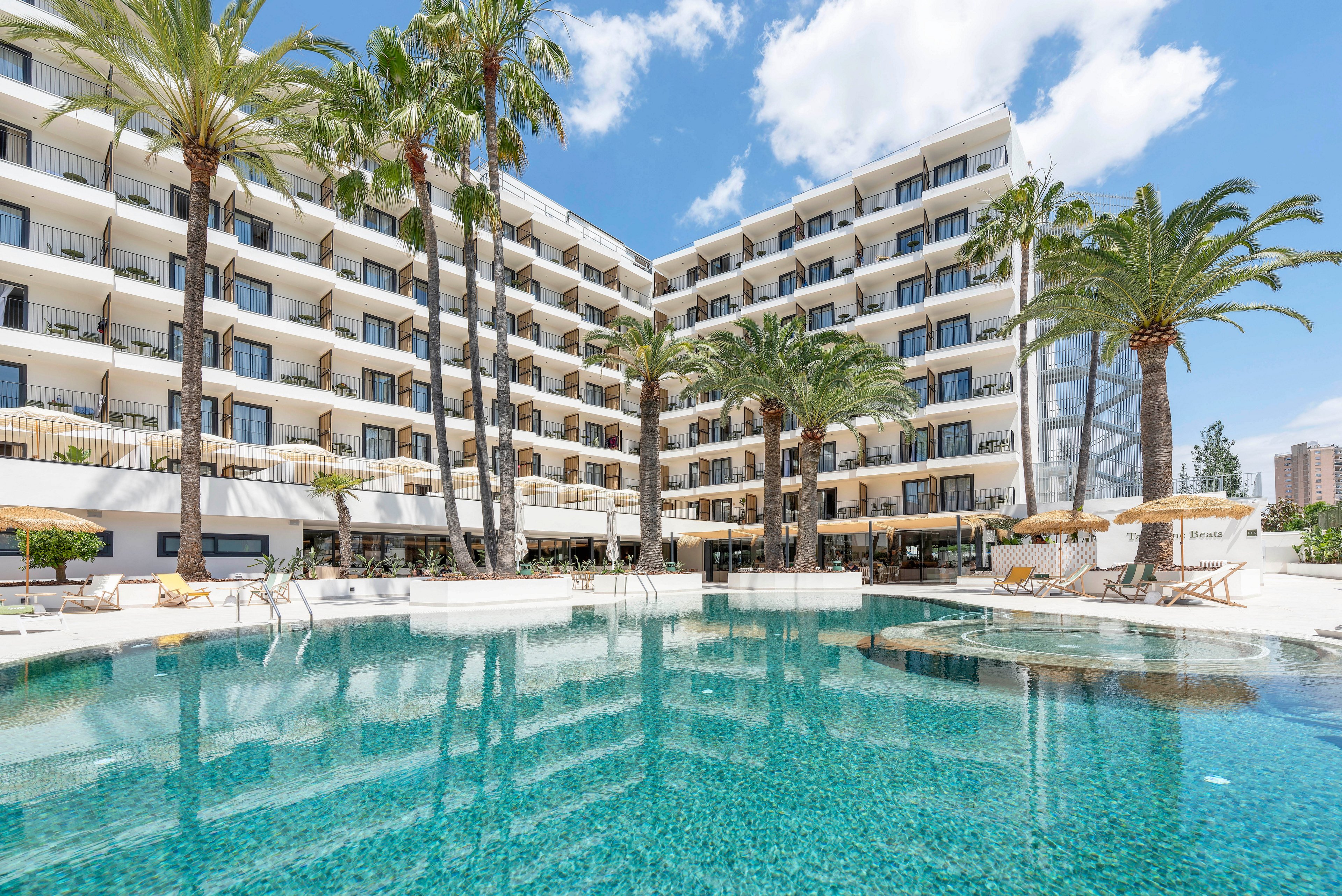 A large modern resort hotel overlooks a clear turquoise swimming pool. Tall palm trees frame the multi-story building with balconies facing the water. Sun loungers and umbrellas are arranged around the poolside deck, suggesting a relaxing vacation setting. The bright blue sky with scattered clouds enhances the sunny, tropical atmosphere.