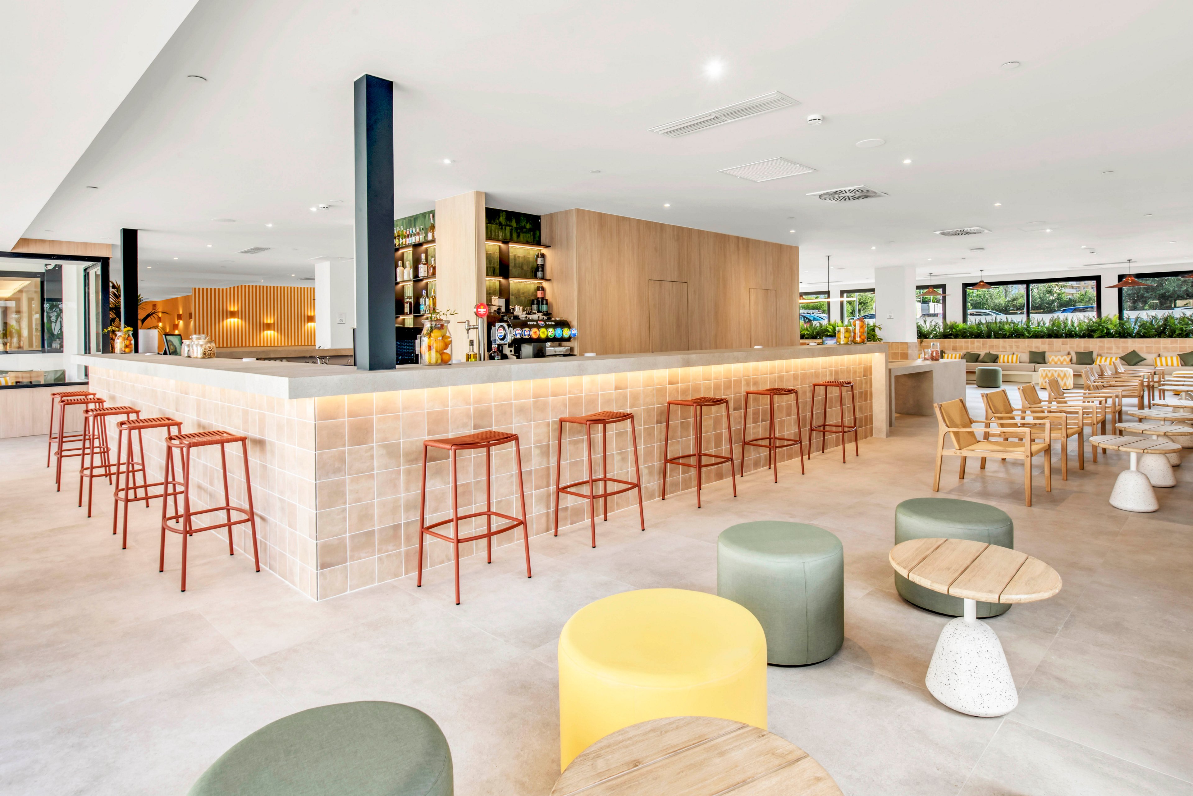 Spacious modern bar interior with tiled counter and high stools is shown in a bright, open-plan setting. The scene features a mix of bar seating and casual dining tables with wooden chairs and low upholstered poufs. Large windows bring in natural light and views of greenery, enhancing the airy atmosphere. Neutral tones, light wood, and soft pastel accents create a clean, minimalist, and welcoming mood.