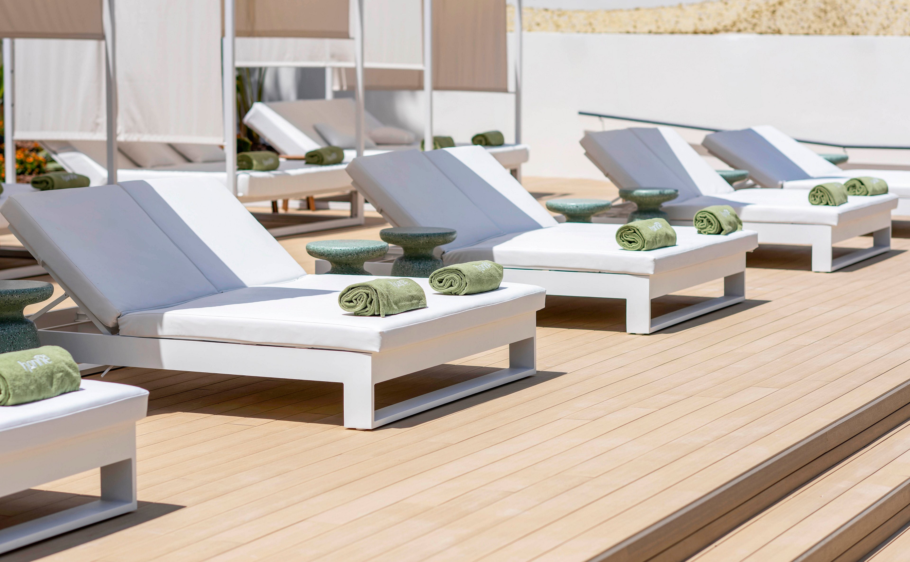 The image shows a neat row of modern white sun loungers arranged on a light wooden deck. Each lounger has a green rolled towel placed on a small side table, suggesting a prepared and welcoming poolside or spa environment. The setting appears bright and sunny, with clean lines and neutral tones creating a minimalist, upscale atmosphere. No people are present, emphasizing relaxation, order, and resort-style comfort.
