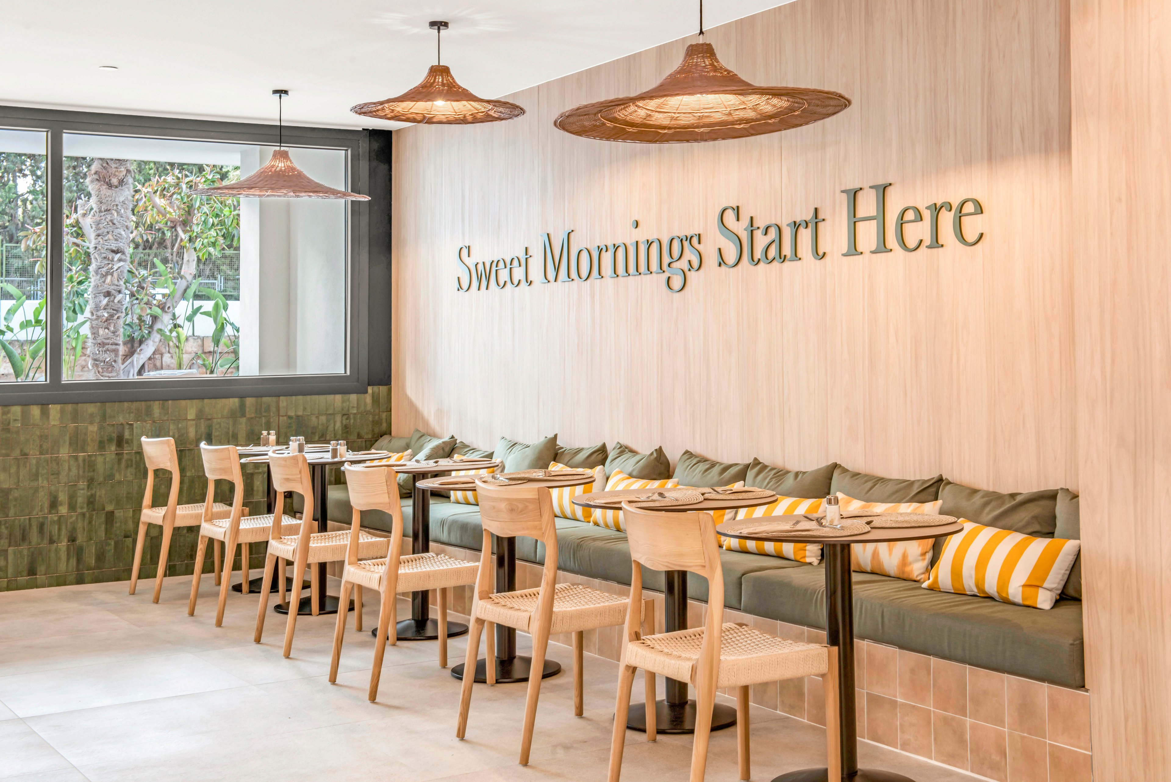 A contemporary café interior features a long cushioned bench with wooden chairs and small tables neatly arranged in a row. The wall displays the phrase "Sweet Mornings Start Here" in large, elegant lettering. Warm pendant lights hang from the ceiling, complementing the light wood paneling and green tile accents. Striped yellow and white pillows add a cheerful touch to the relaxed, inviting atmosphere.