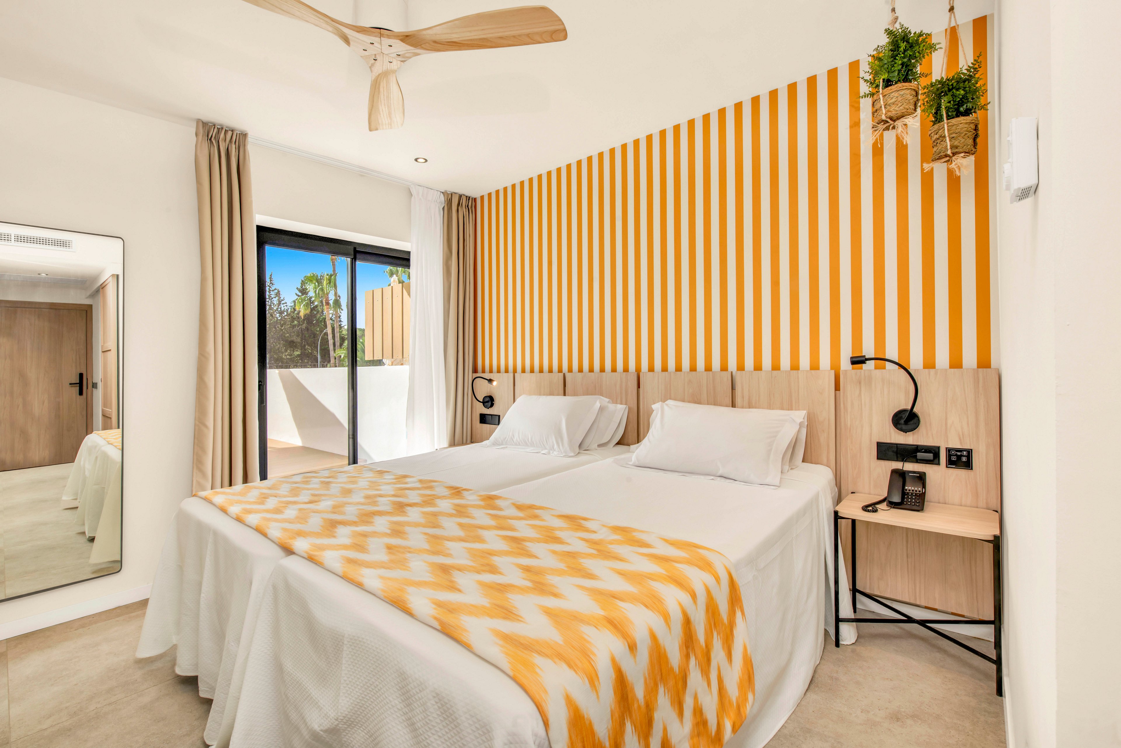 Bright contemporary bedroom featuring two twin beds with white linens and a yellow chevron blanket. The room has a bold yellow and white striped accent wall behind the headboard and light wood furniture. A large sliding glass door opens to an outdoor terrace, bringing in natural light. Neutral curtains, a ceiling fan, hanging plants, and a wall mirror complete the cozy, modern decor.