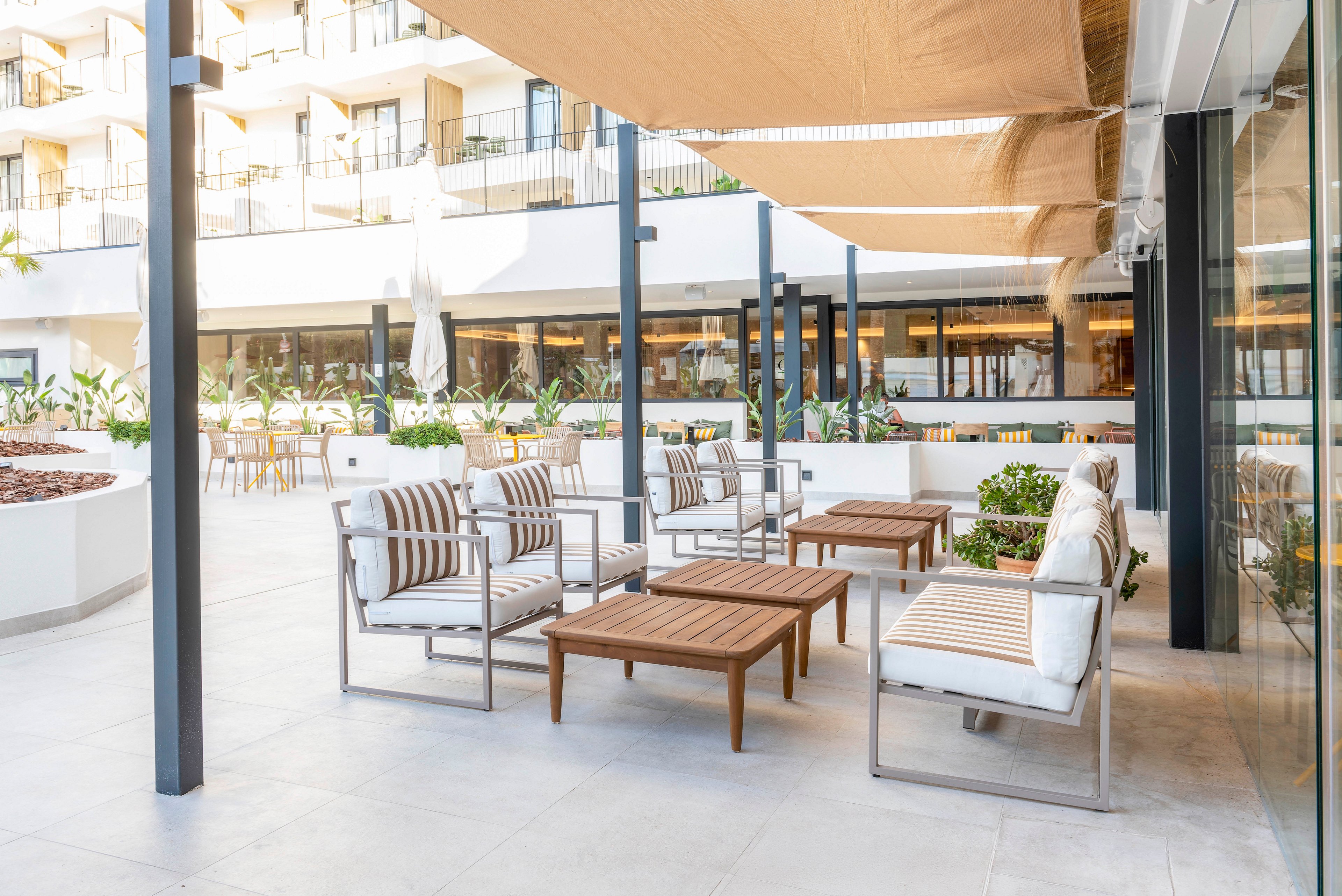 Contemporary outdoor lounge area on a hotel terrace features cushioned chairs and wooden coffee tables. Neutral striped upholstery and light-colored flooring create a bright, airy atmosphere. Shade sails overhead and potted greenery add comfort and a relaxed resort feel. In the background, balconies and an open dining area suggest a vacation or hospitality setting.