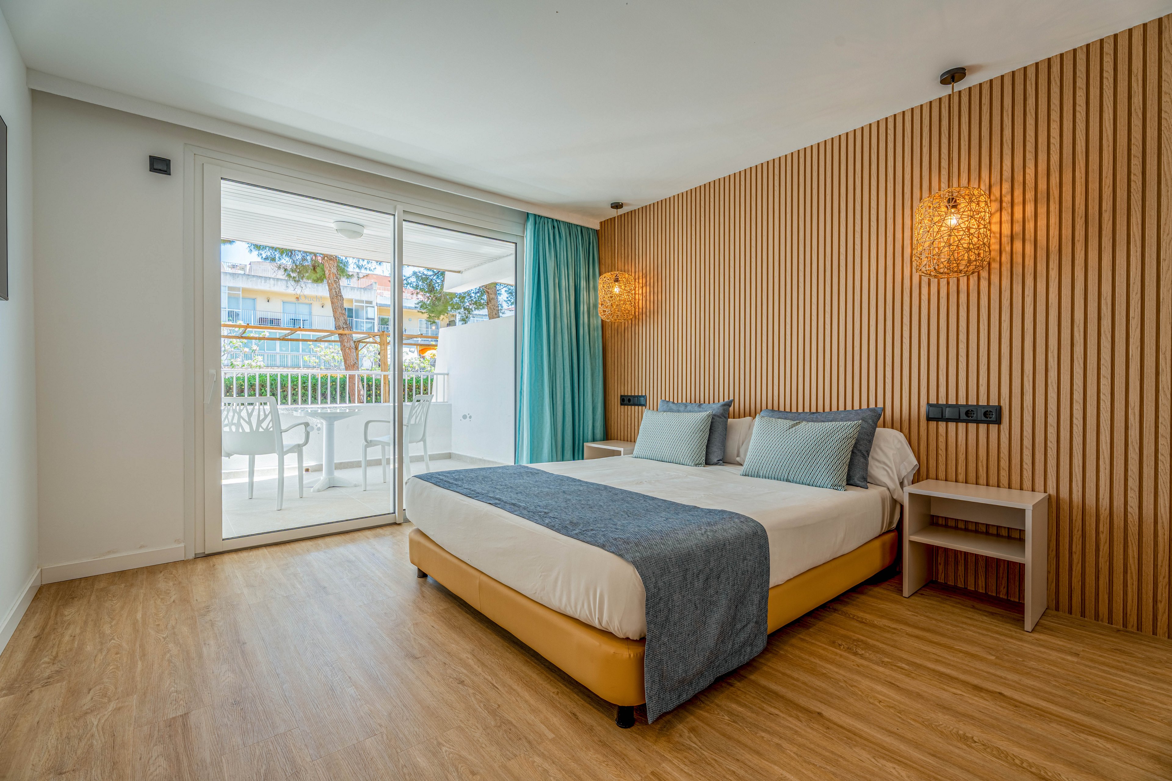A contemporary hotel bedroom features a neatly made double bed with blue and gray accents. The room has a warm wooden floor and a vertical wood slat accent wall with woven pendant lights on each side of the bed. Large sliding glass doors open to a furnished balcony with white chairs and a table, overlooking other buildings and trees. Natural light fills the space, highlighting the clean, minimalist decor and relaxing atmosphere.