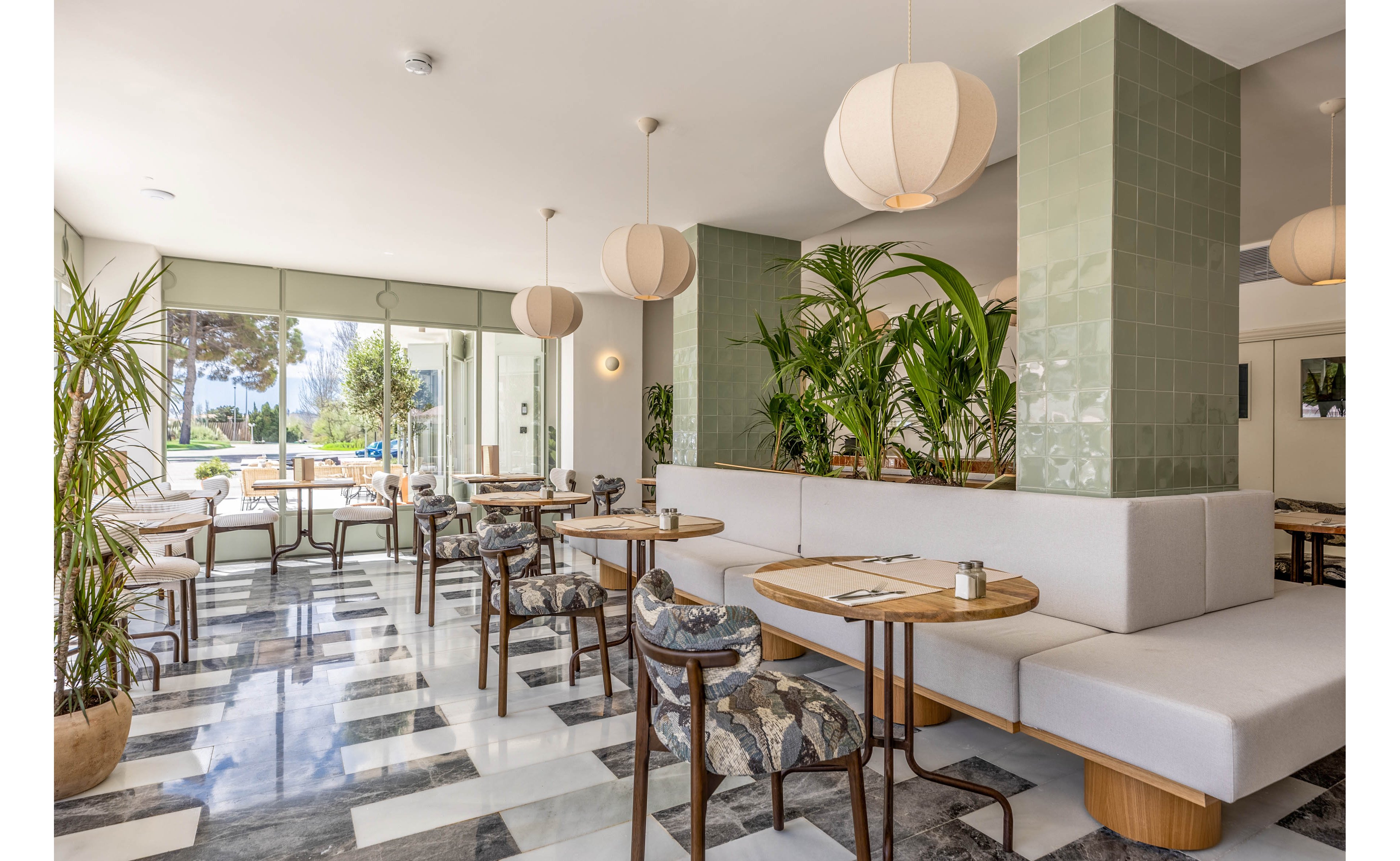 A stylish café interior features light-colored banquette seating and round wooden tables. Large windows fill the space with natural light and offer views of trees and an outdoor area. Pendant lamps hang from the ceiling, while green tiled columns and numerous potted plants add a fresh, organic touch. The black-and-white patterned floor and patterned chair upholstery create a contemporary yet cozy atmosphere.