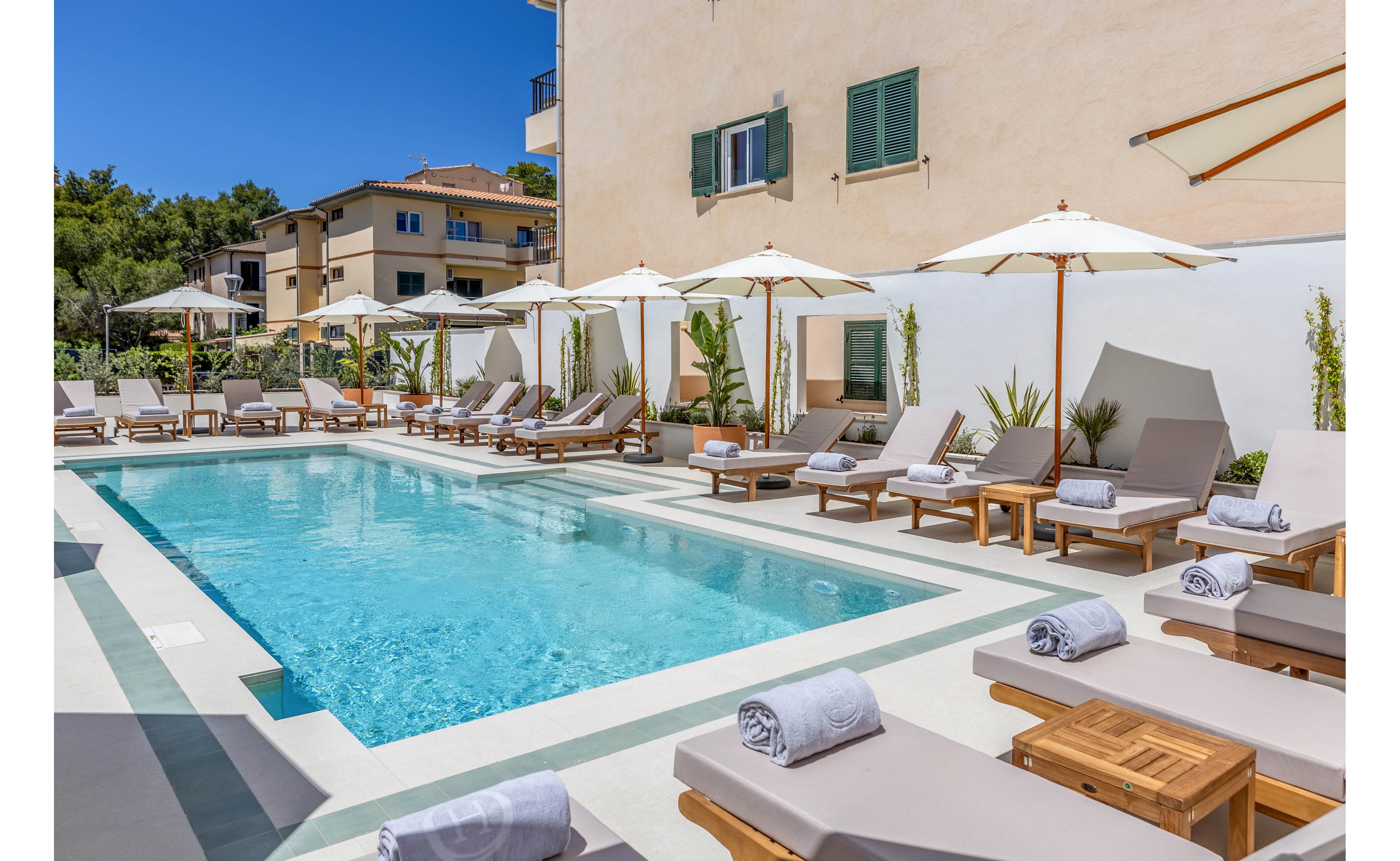 An outdoor swimming pool is surrounded by cushioned sun loungers and white umbrellas at a boutique-style hotel. Rolled towels rest on the loungers and small wooden side tables, suggesting a relaxing resort atmosphere. The clear blue water reflects the bright sky, while potted plants add greenery along the white walls. Nearby residential buildings and trees frame the tranquil courtyard setting.