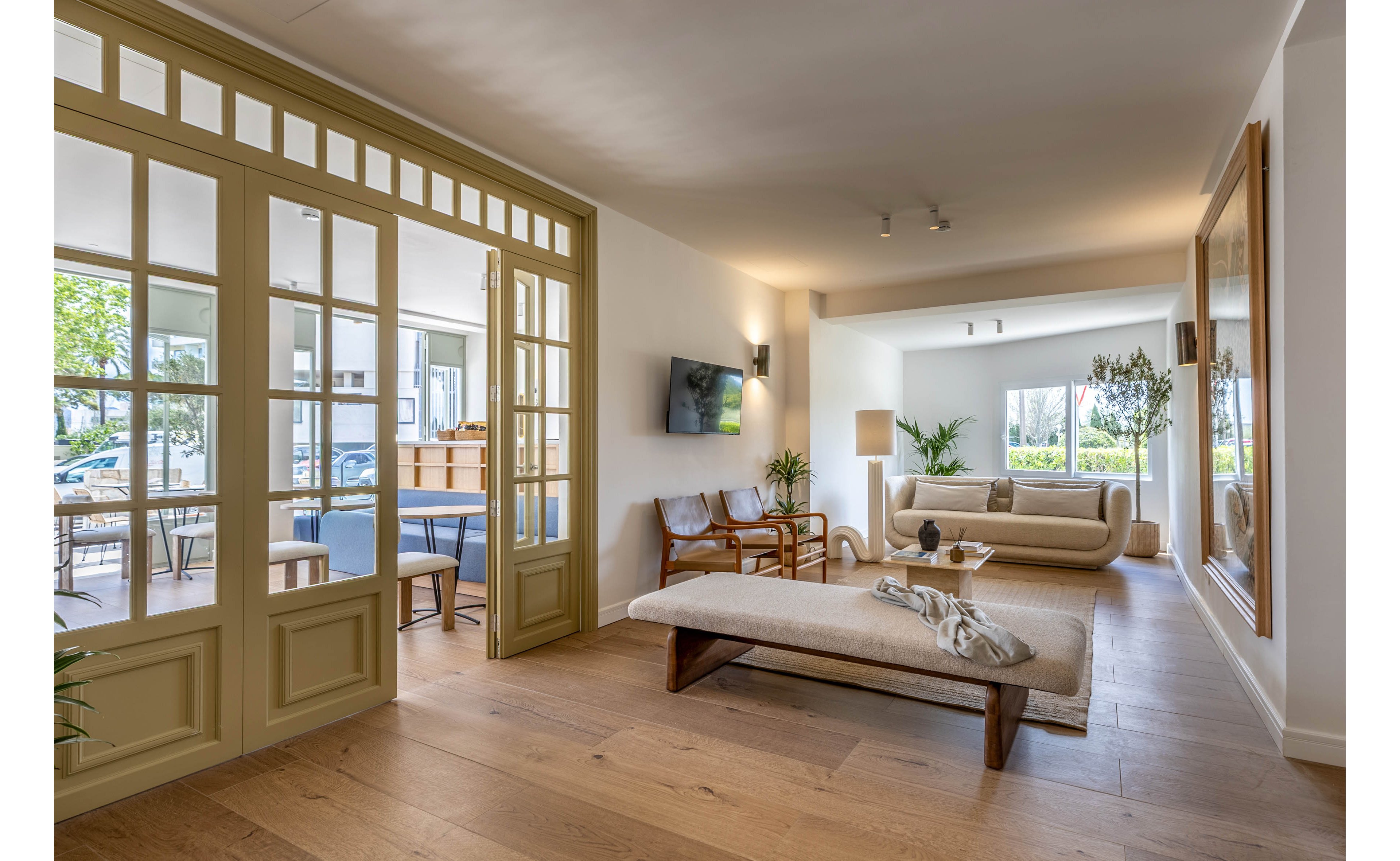 Spacious modern living room featuring a low bench-style daybed and a neutral sofa. A glass-paneled partition with wooden framing separates the lounge from a dining or meeting area with blue chairs. Large windows bring in natural light and views of greenery, while indoor plants and minimalist decor create a calm atmosphere. A wall-mounted television and simple lighting fixtures complete the contemporary interior design.