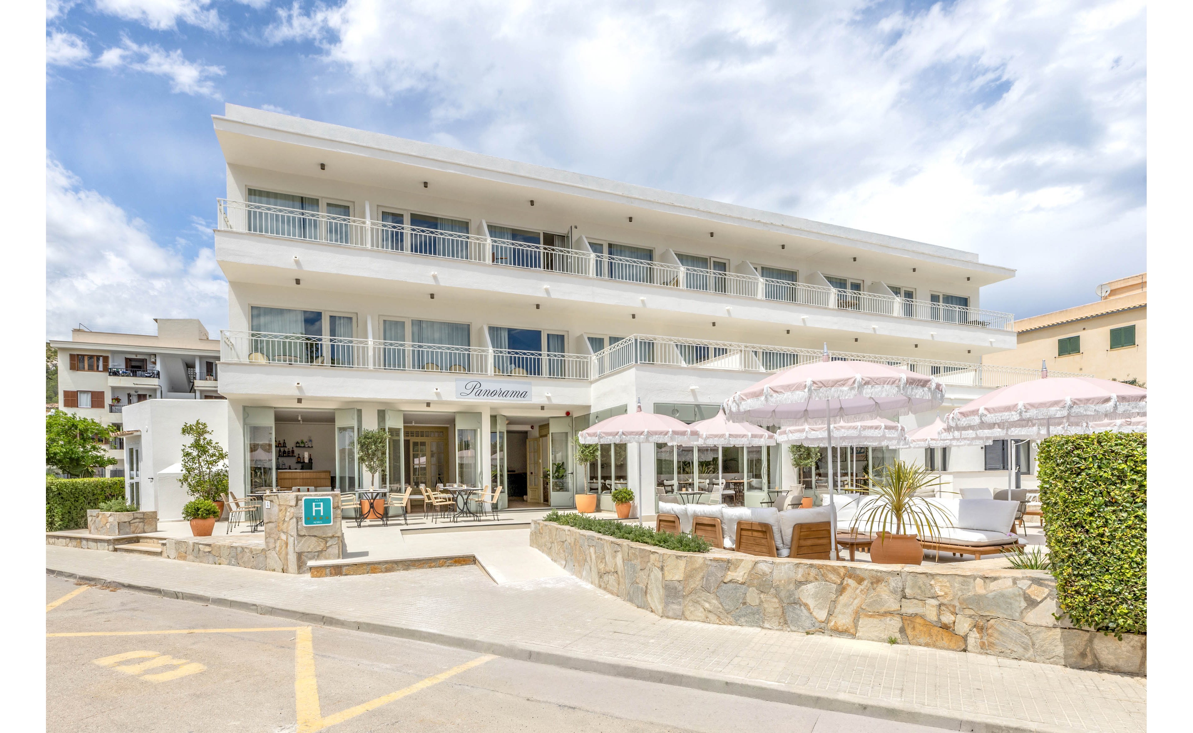 A contemporary white hotel building stands on a sunny street corner with multiple balconies and large windows. The ground floor features an open-air terrace with tables, chairs, and pink parasols arranged for guests. Potted plants and trimmed hedges add greenery along the stone wall and entrance area. The bright daylight and partly cloudy sky create a relaxed, inviting vacation atmosphere.