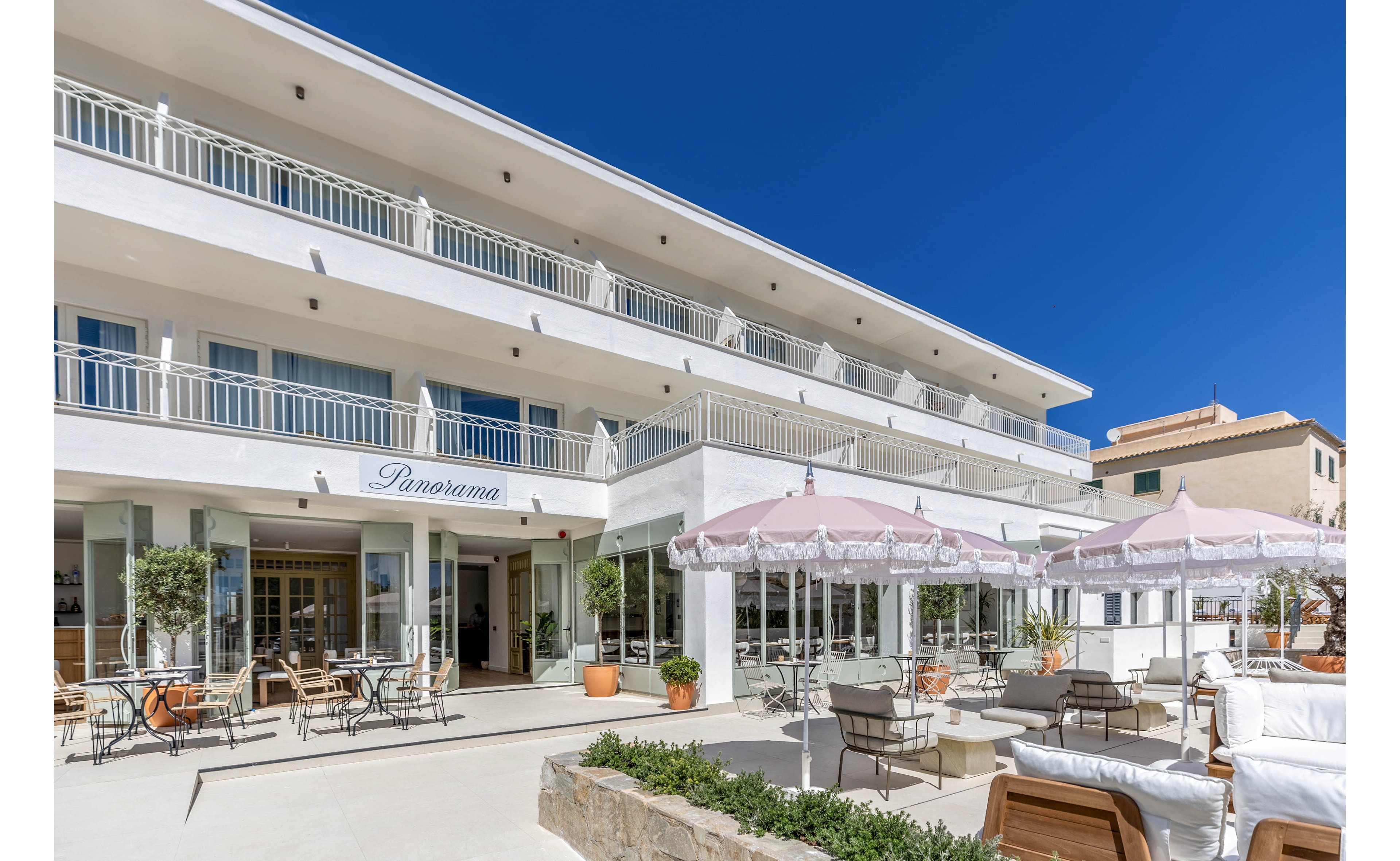 A bright, modern hotel named Panorama features multiple stories with white balconies and large windows. In the foreground, an outdoor terrace is furnished with tables, chairs, and cushioned lounge seating under pale pink umbrellas. Potted plants and small trees add greenery to the clean, minimalist architecture. The scene is lit by clear blue skies, suggesting a warm, inviting coastal destination.