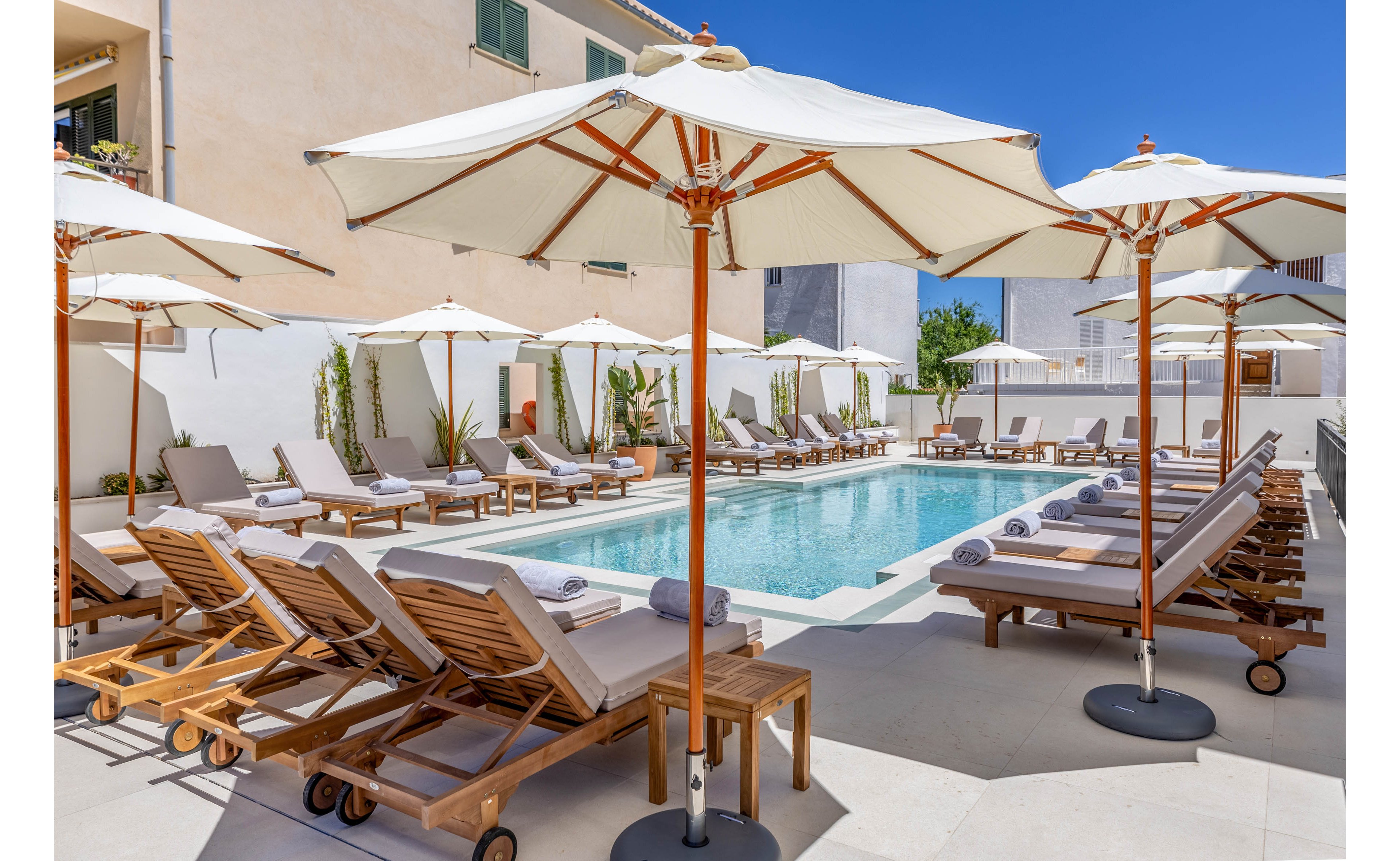 An outdoor hotel swimming pool is surrounded by rows of wooden sun loungers with neatly rolled towels. Large white umbrellas provide shade along the pool deck, creating a resort-style atmosphere. The setting is bright and sunny, with clear blue water reflecting the sky. Modern buildings and white walls frame the tranquil courtyard space.