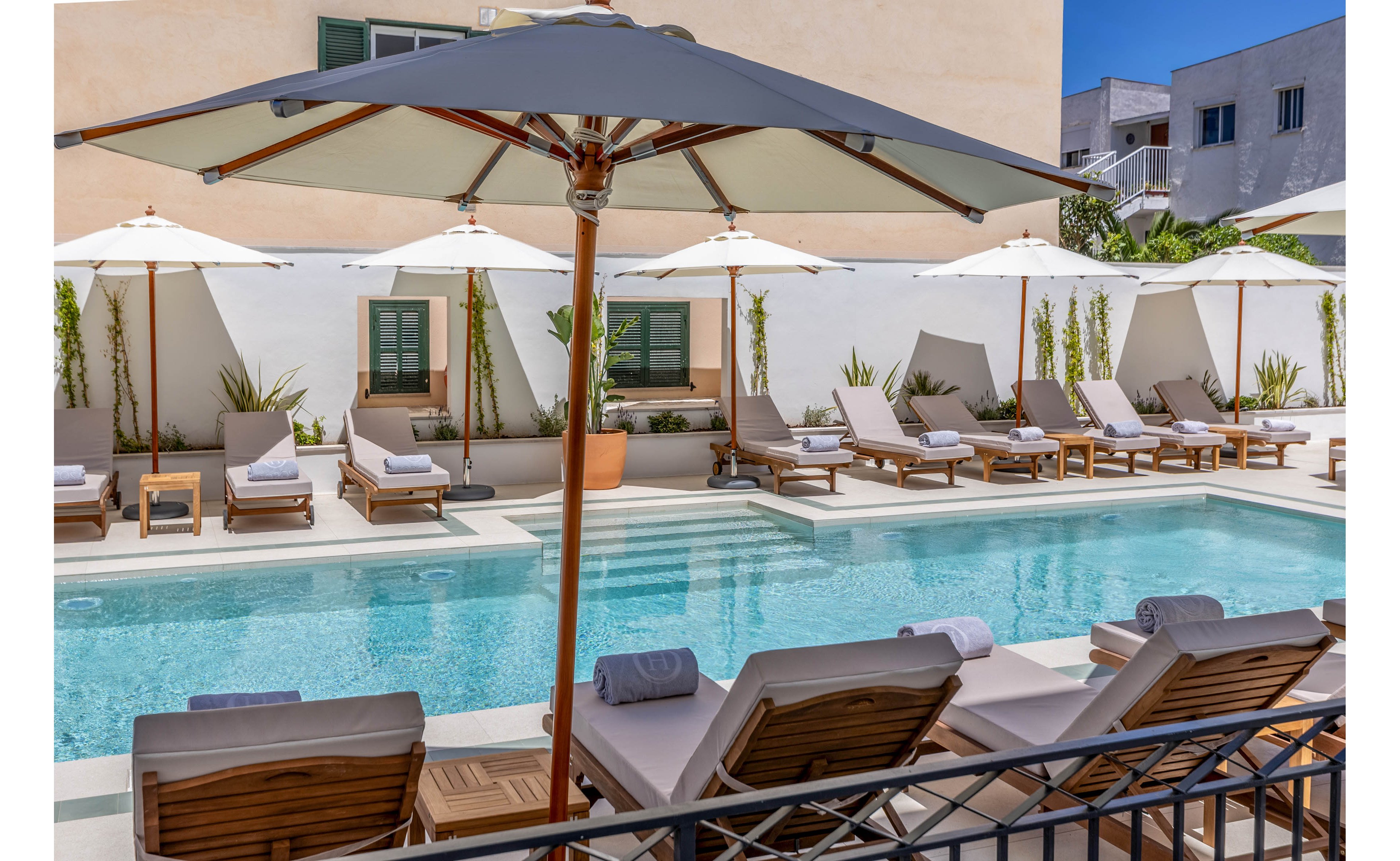 An inviting outdoor swimming pool is surrounded by cushioned wooden loungers and large white umbrellas. Rolled towels rest on each chair, suggesting a prepared and relaxing poolside experience. The setting appears to be a modern boutique hotel courtyard with white walls, potted plants, and minimal architectural details. Bright daylight and clear blue water create a calm, luxurious vacation atmosphere.