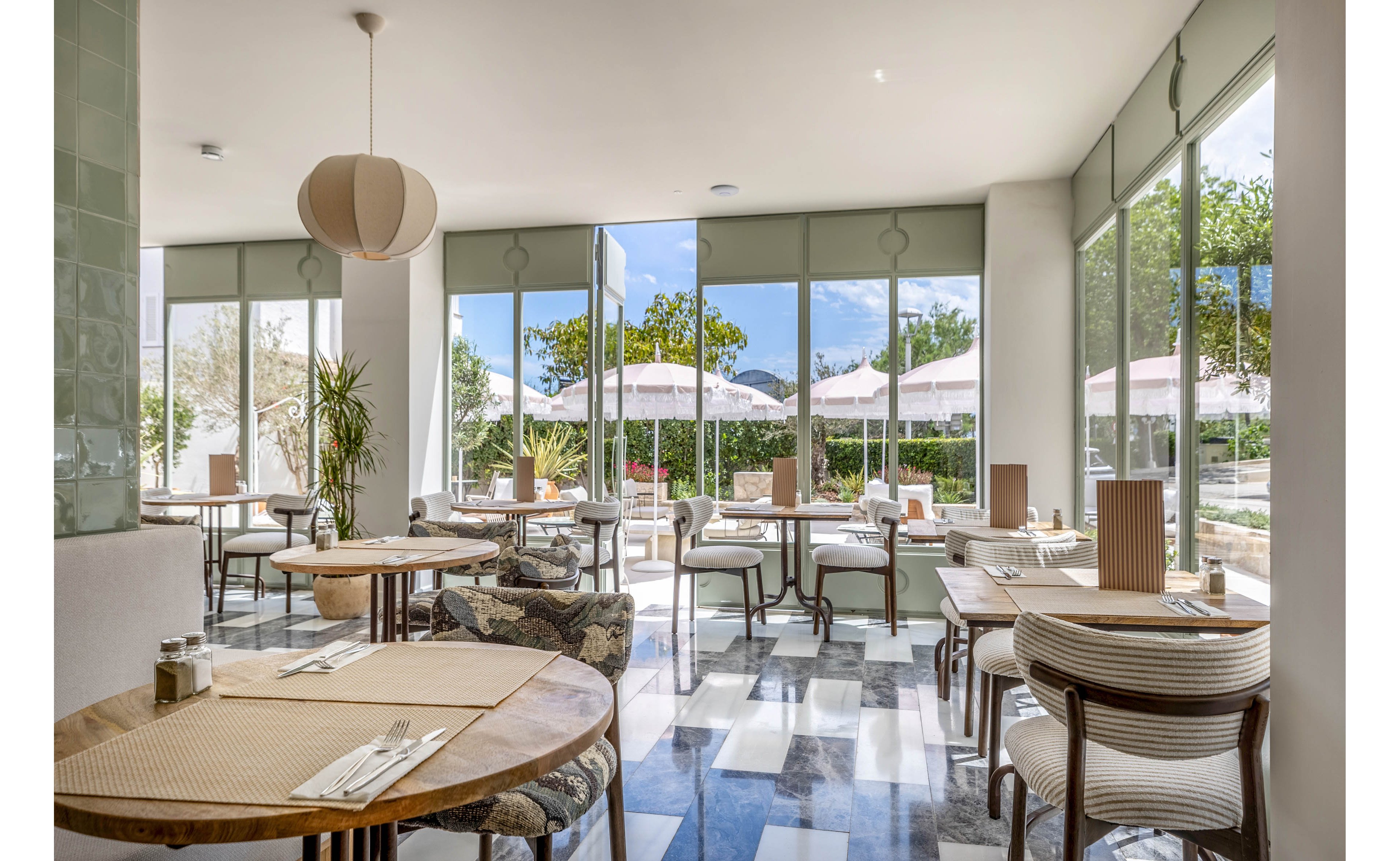Sunlit restaurant interior features neatly arranged round and square tables with woven placemats and simple table settings. Large floor-to-ceiling windows open onto an outdoor patio with umbrellas and greenery. Soft neutral tones, light wood furniture, and patterned chairs create a relaxed, contemporary atmosphere. The space appears calm and empty, ready to welcome guests for a meal.