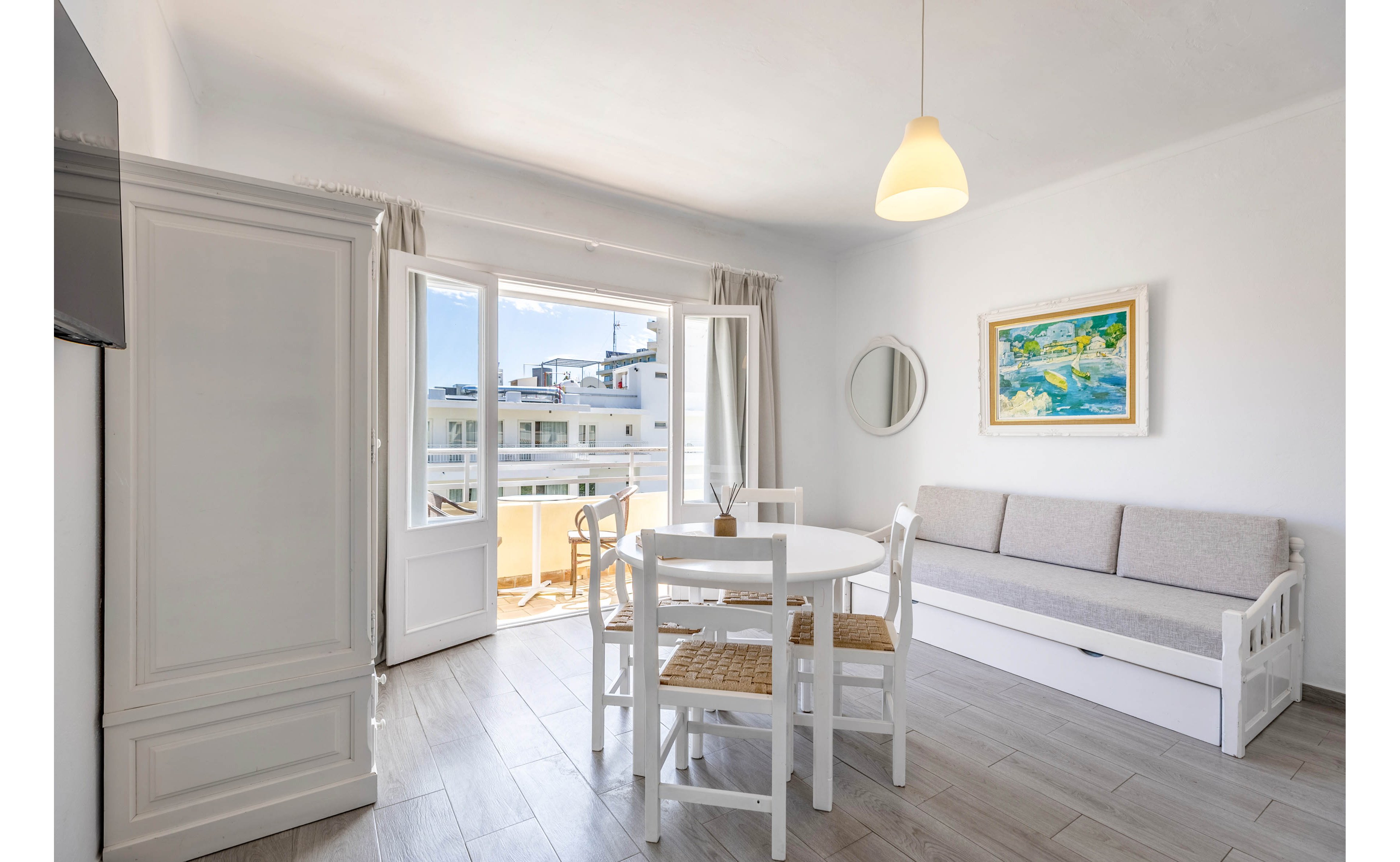 Sunlit living and dining room in a coastal apartment with white furniture and neutral decor. A round table with four chairs sits in the center of the room beside a light gray sofa. Large glass doors open to a balcony with views of neighboring white buildings. Soft natural light, pale wood flooring, and a colorful wall painting create an airy, relaxed atmosphere.