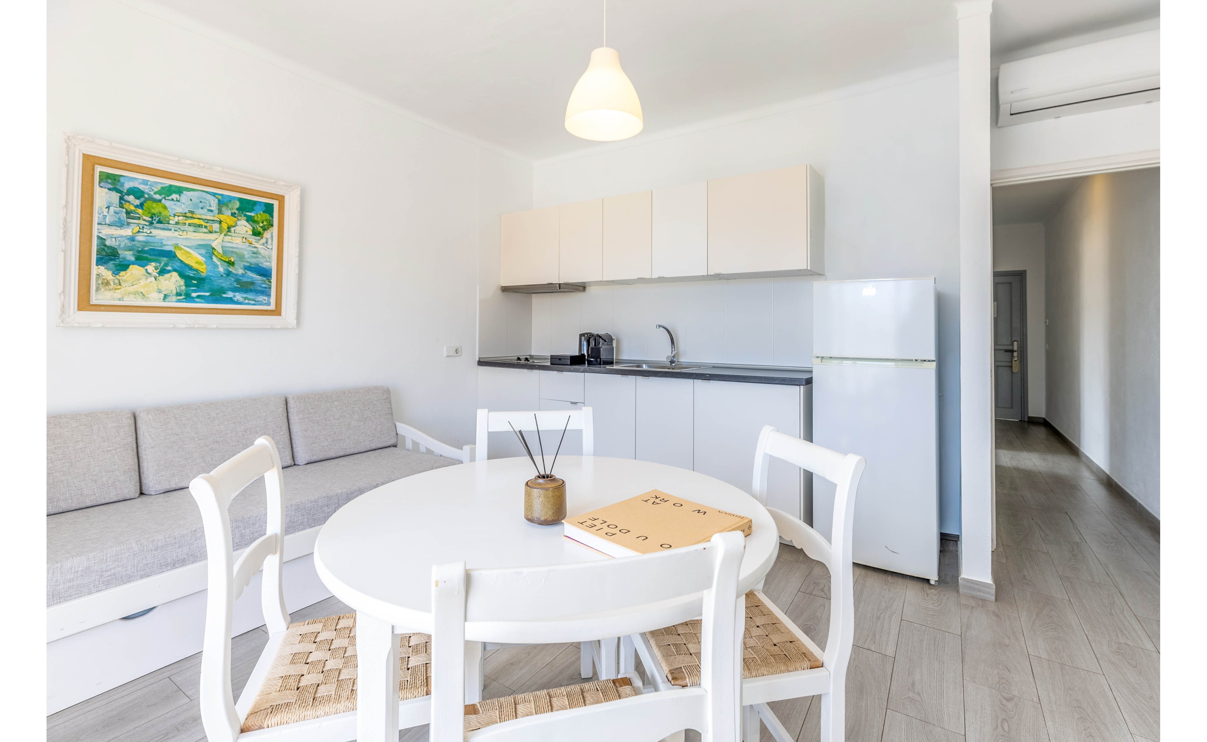 A compact studio apartment features a small open kitchen and dining space. A round white table with woven chairs sits in the foreground, with a neutral sofa and colorful wall art on the left. The kitchenette includes upper cabinets, a sink, countertop appliances, and a tall white refrigerator. Light wood flooring and white walls create an airy, minimalist atmosphere, enhanced by a hanging pendant light and a long hallway leading to other rooms.