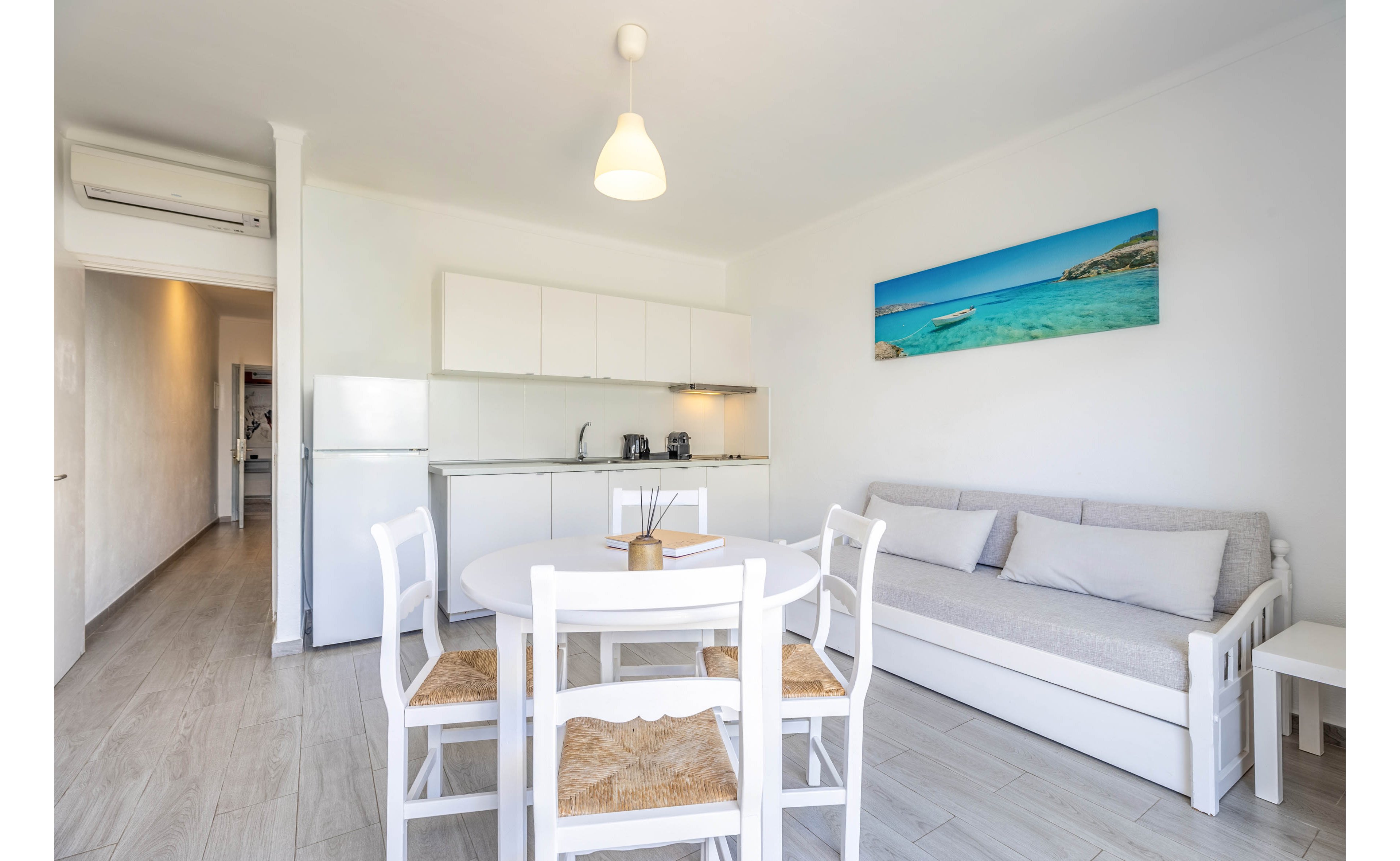 A bright studio apartment features a compact white kitchen and a small dining table with four chairs. A light gray sofa with cushions sits against the wall beside a white side table. The space is decorated in a minimalist style with neutral tones and a single colorful seascape artwork on the wall. Natural light and pale flooring enhance the airy, modern feel of the interior.