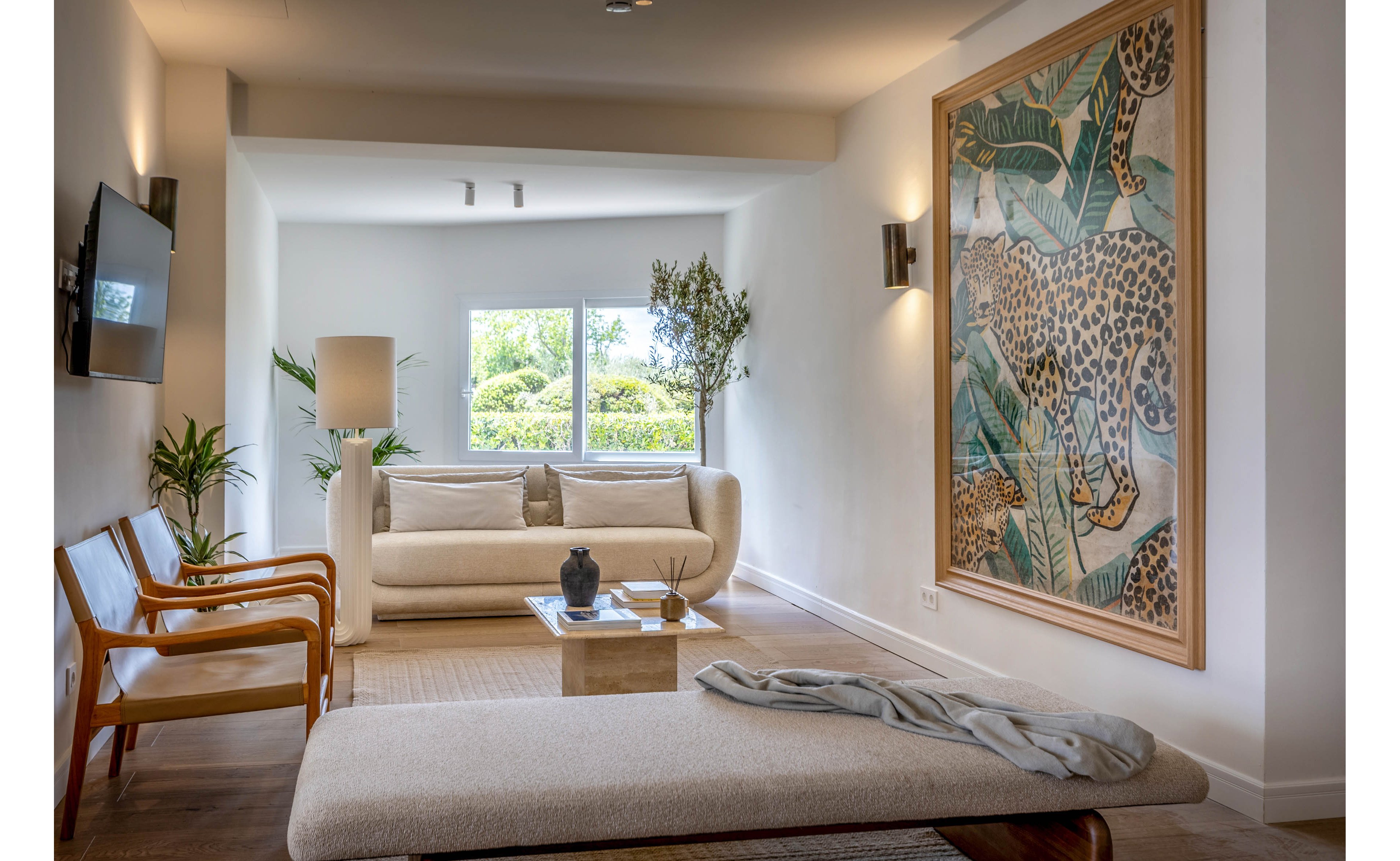 A contemporary living room features a light-colored sofa and a cushioned bench facing a low coffee table. Large windows at the back bring in natural light and views of greenery outside. A prominent framed artwork of a leopard among tropical leaves decorates the right wall, adding a bold focal point. Neutral tones, wooden chairs, and indoor plants create a calm, minimalist atmosphere.
