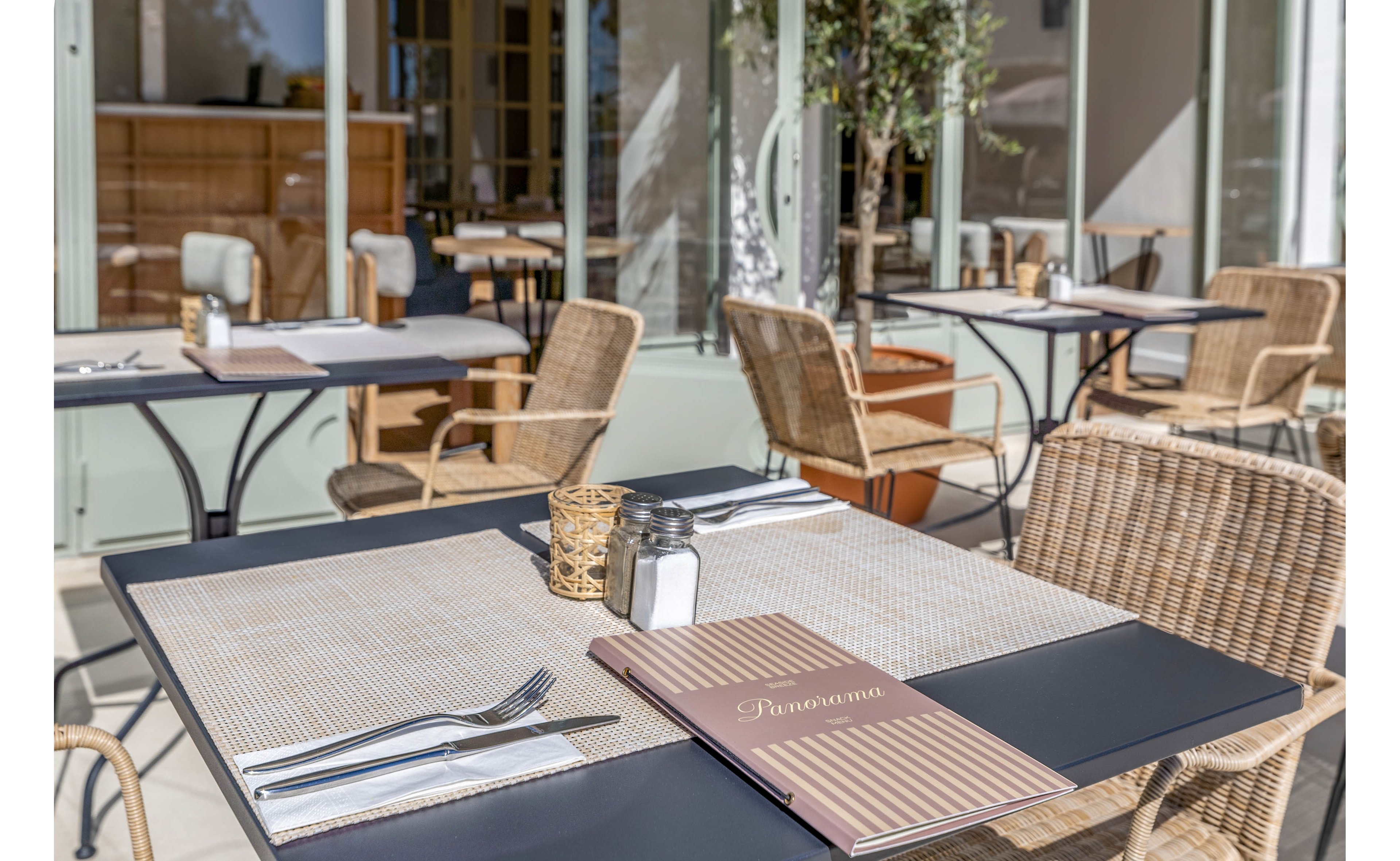 An outdoor café terrace features neatly set tables with woven placemats and cutlery. A closed menu labeled "Panorama" rests on the foreground table beside salt and pepper shakers. Wicker chairs surround the dark tabletops, creating a relaxed dining atmosphere. Large glass doors and soft natural light suggest a bright, inviting setting for guests.