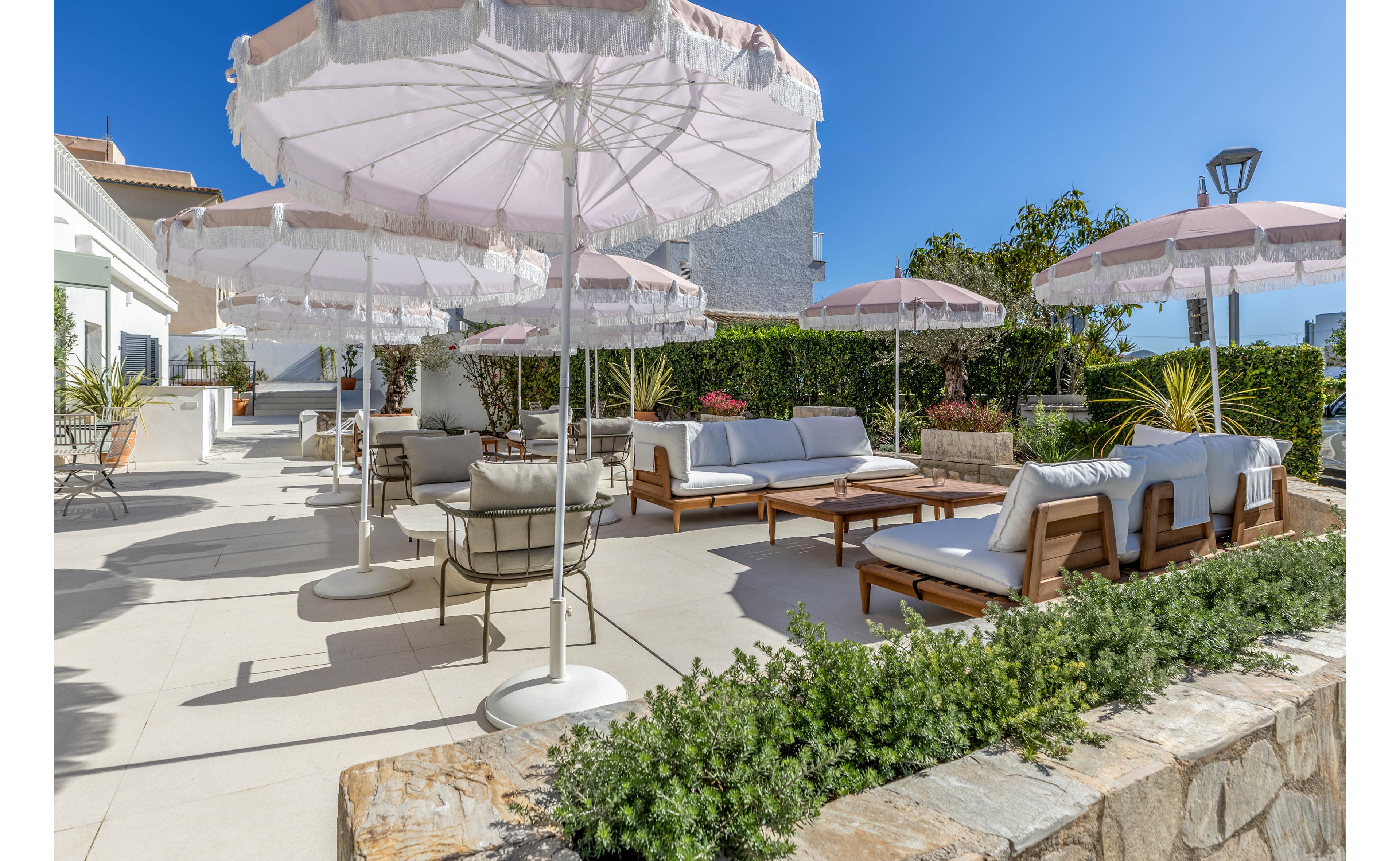 An elegant outdoor lounge area features white cushioned sofas and chairs arranged on a spacious terrace. Large pale pink umbrellas provide shade under a clear blue sky. Green shrubs and potted plants border the stone and tile surfaces, adding a touch of nature. The bright sunlight and clean design create a relaxed, resort-like atmosphere.