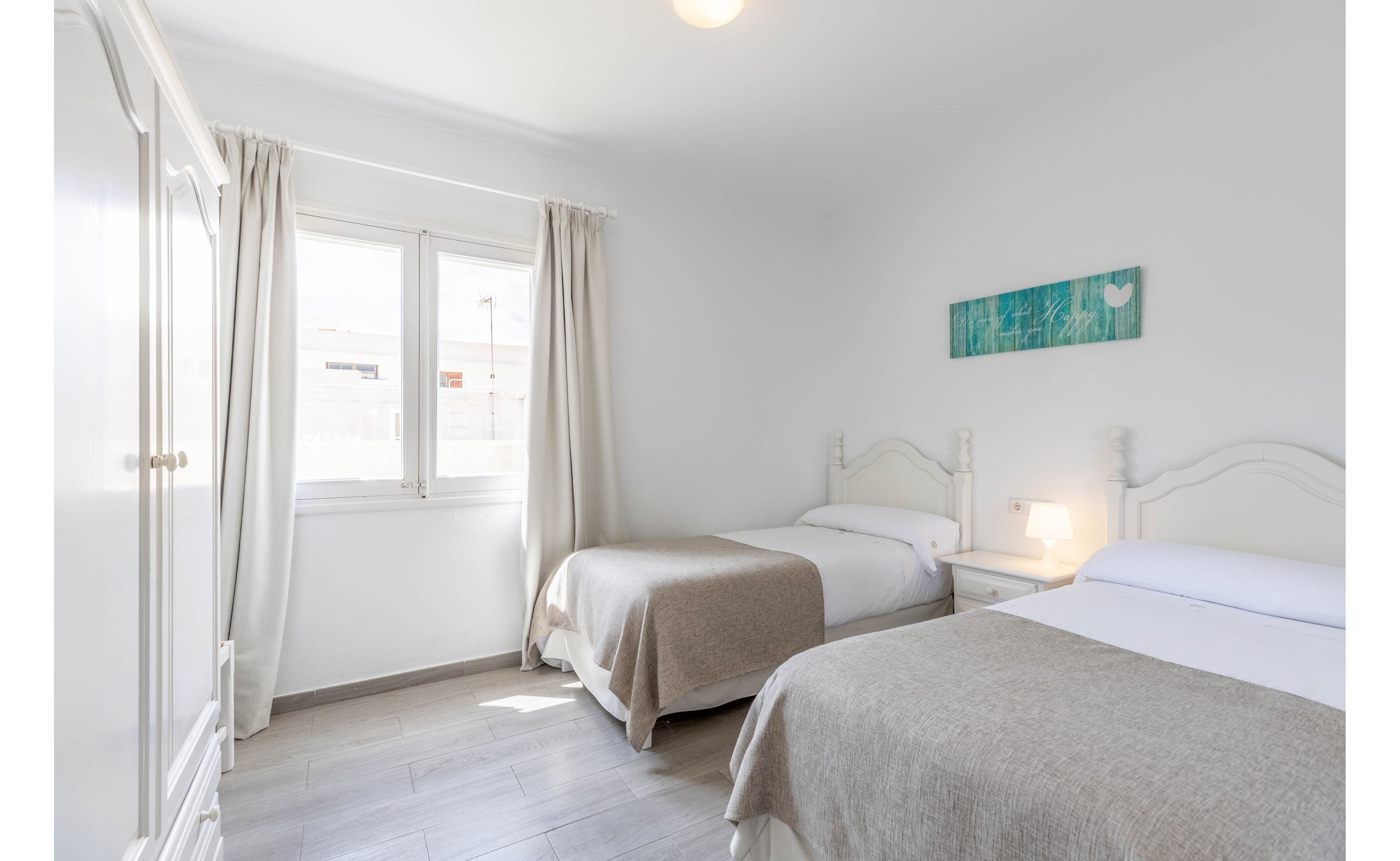 A bright twin bedroom features two single beds with white linens and beige throws. Natural light streams in through a large window with light curtains, illuminating the white walls and simple furnishings. A small bedside table with a lit lamp sits between the beds, adding warmth to the space. A turquoise wall art piece provides a subtle accent in the otherwise neutral, minimalist room.