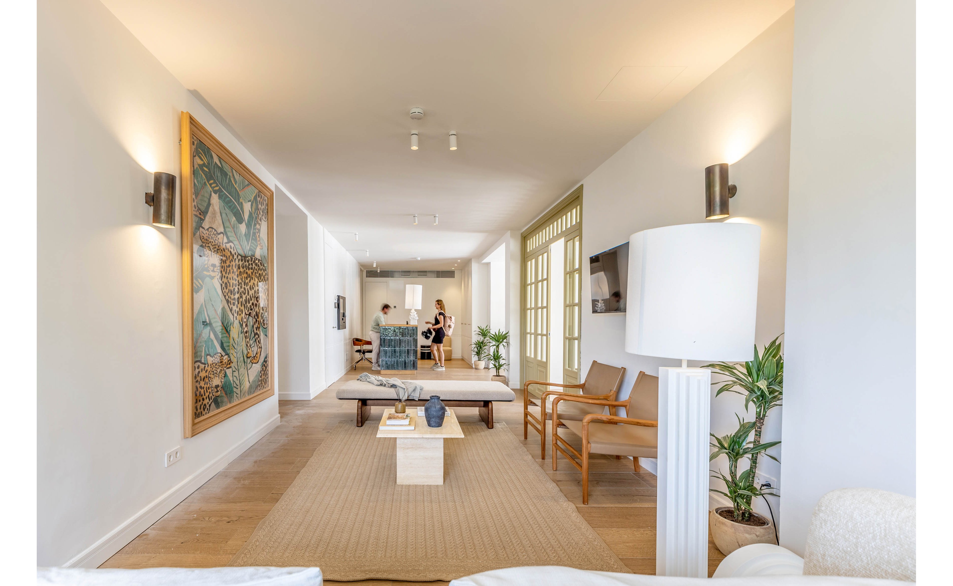 A spacious modern living room features neutral furnishings and minimalist decor. A large artwork hangs on the wall and contemporary chairs and benches are arranged around a low coffee table. Soft wall lighting and a tall floor lamp create a warm, inviting atmosphere. In the background, a small group of adults stands near a dining area, suggesting a relaxed social gathering.