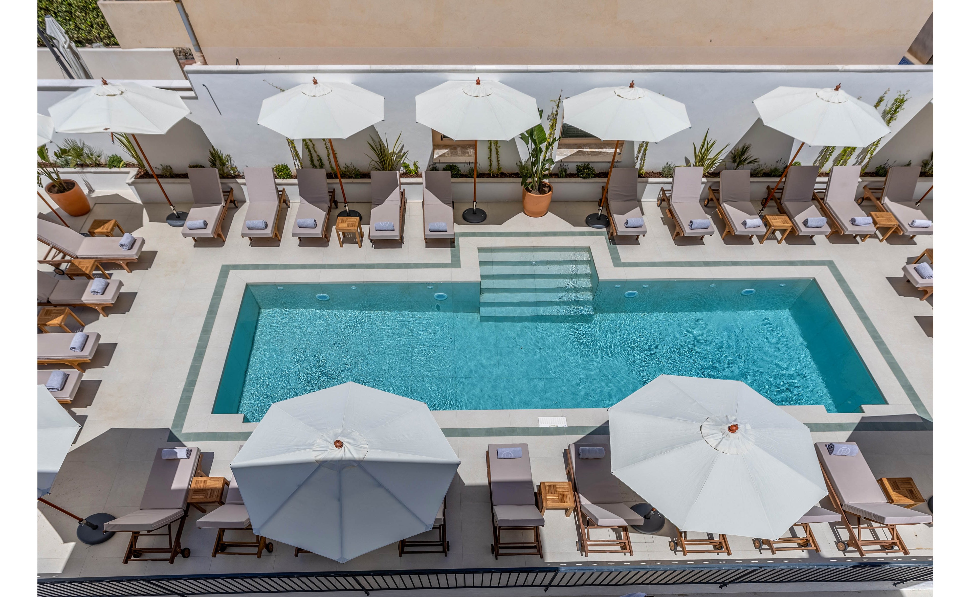 An elevated view shows a rectangular outdoor swimming pool surrounded by neatly arranged sun loungers. Large white umbrellas provide shade along both sides of the pool, creating a symmetrical and orderly layout. The setting appears to be a modern rooftop or terrace area with light-colored tiles and potted plants along the back wall. The overall atmosphere is calm, minimalistic, and designed for relaxation in a resort or hotel environment.