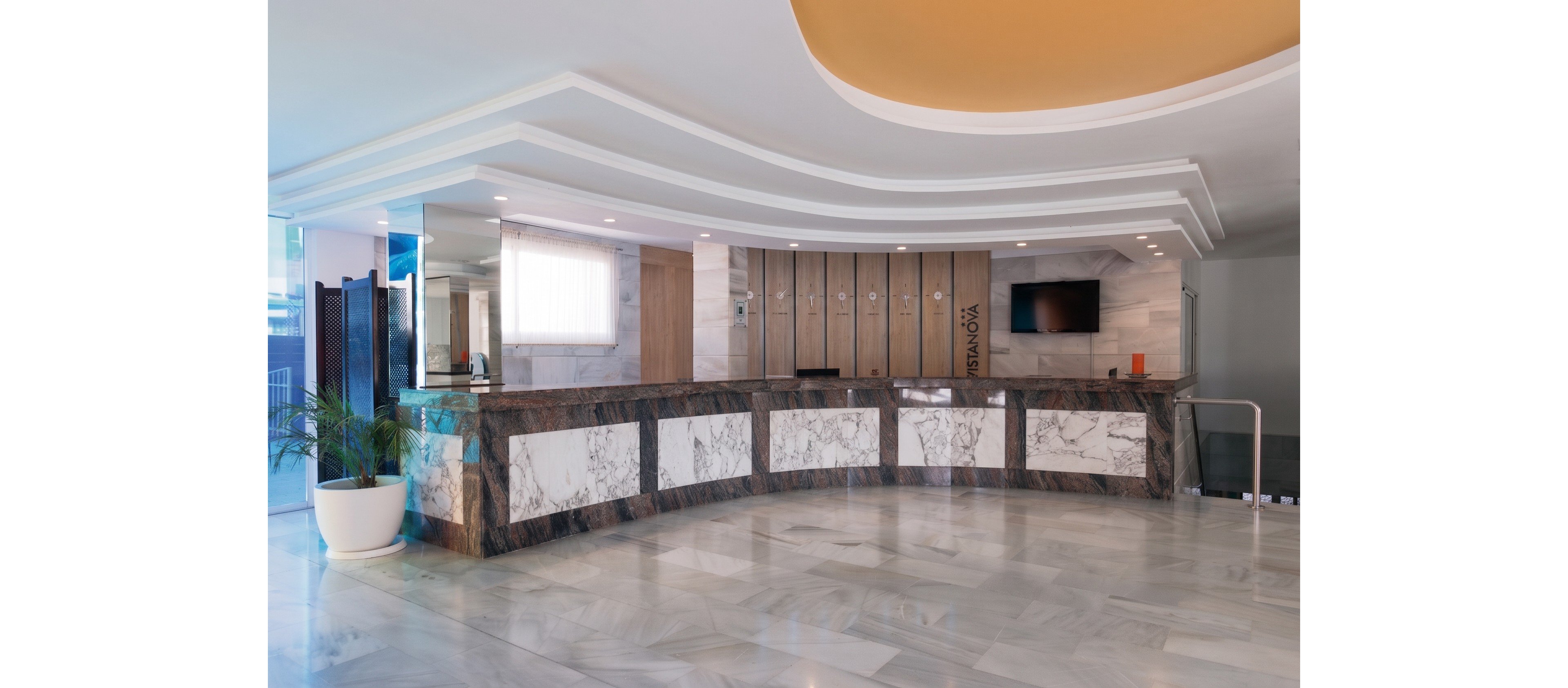 Spacious hotel reception area featuring a long marble-front desk dominates the scene. The lobby has a polished stone floor, contemporary ceiling design, and wood-paneled wall behind the counter. A flat-screen television and small decorative items sit on the back counter, while a potted plant adds greenery near the entrance. The overall atmosphere is clean, modern, and welcoming, with soft natural and artificial lighting.