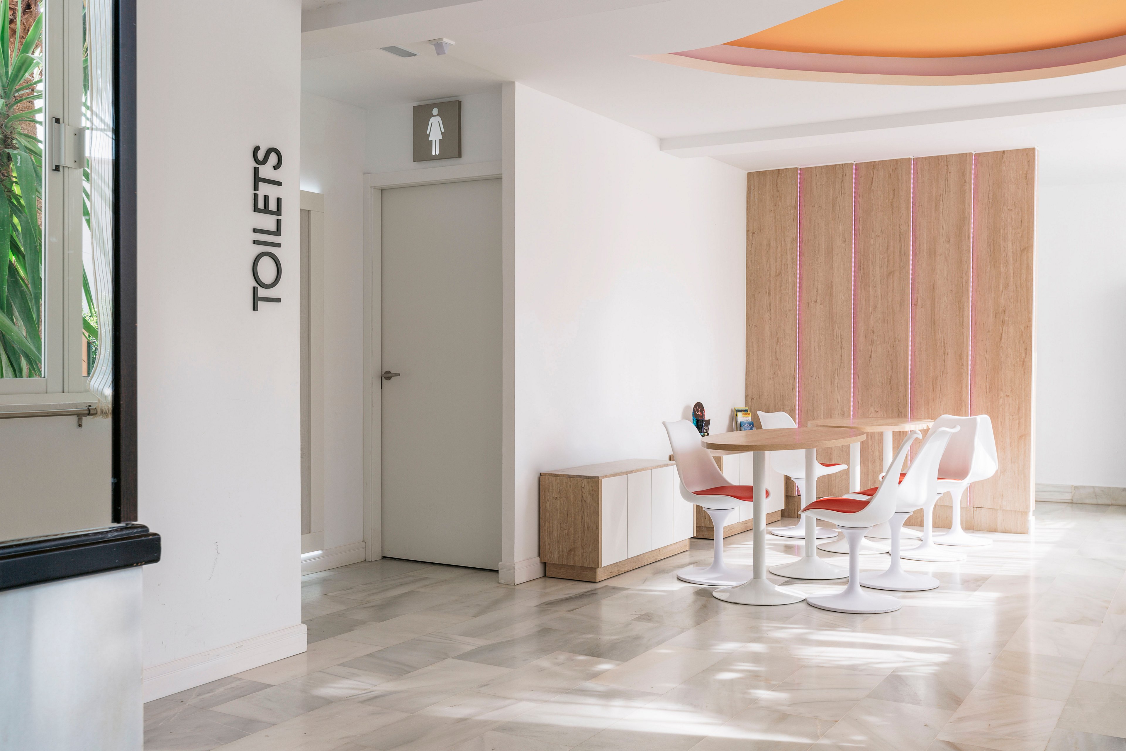 A clean, modern interior hallway features a white door marked as a toilet entrance. Nearby, a small seating area with white chairs and a round table sits against a light wood wall with vertical pink accents. The floor is finished in light marble or tile, and natural light streams in from the left side. The overall space feels bright, minimal, and contemporary.
