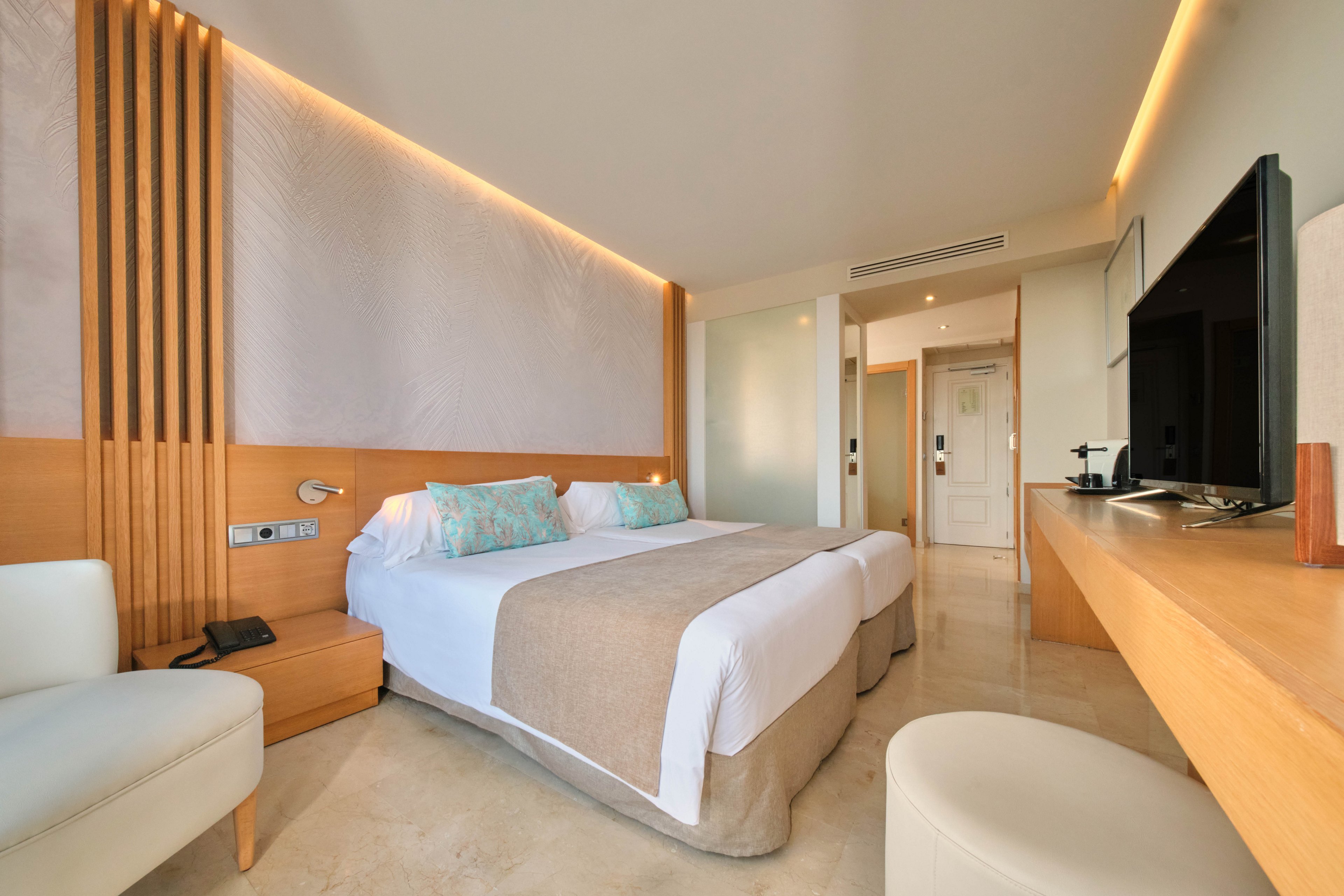 A contemporary hotel room featuring two neatly arranged twin beds with beige and white bedding. The room is well-lit with warm lighting and includes wooden accents, a flat-screen TV, and a desk. The decor is minimalistic with soft pastel tones and a clean, inviting atmosphere.