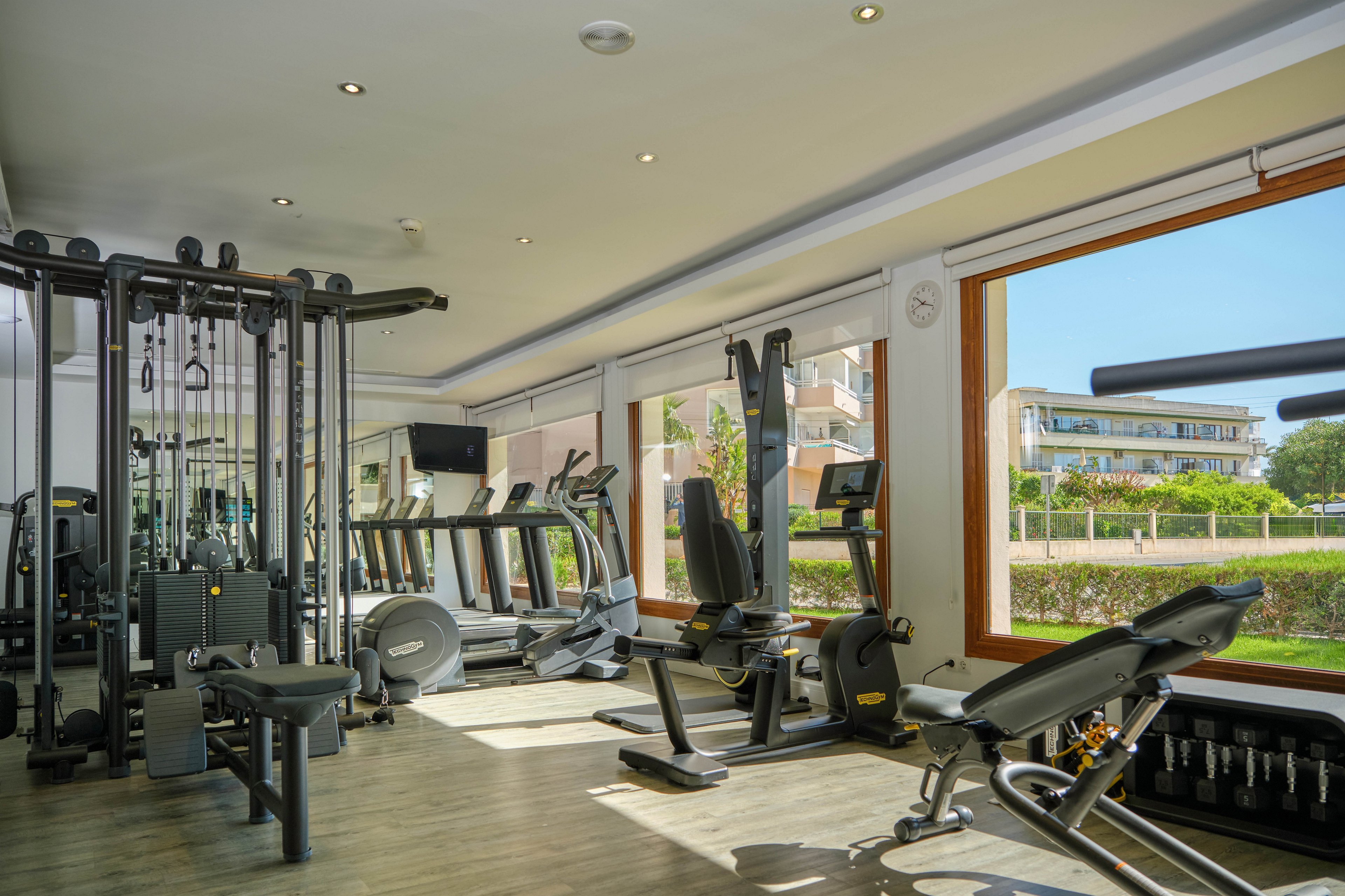 A bright, modern fitness room features various strength and cardio machines neatly arranged. Large windows line one side of the gym, offering views of greenery and nearby buildings. The space has light wood flooring, neutral walls, and recessed ceiling lights that create a clean, inviting atmosphere. No people are present, emphasizing the equipment and open workout area.