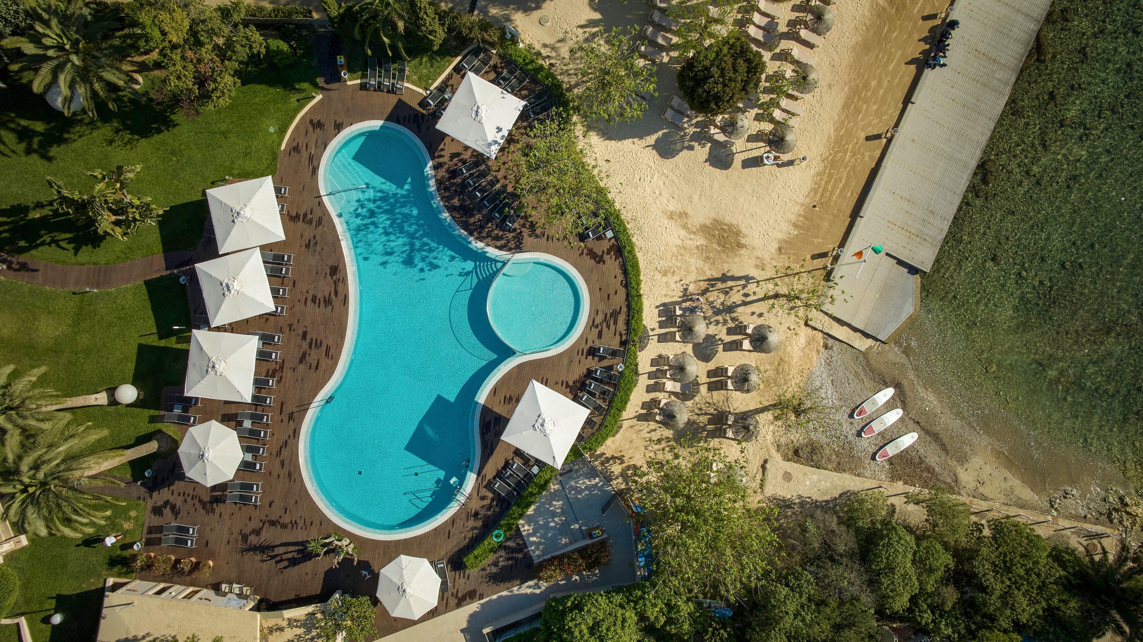 An aerial view shows a curving resort swimming pool surrounded by lounge chairs and large white umbrellas. The pool area is bordered by wooden decking and lush green landscaping. To the side, a sandy beach with additional loungers leads down to the water. Several small boats or paddleboards are lined up near the shoreline, emphasizing the coastal vacation setting.