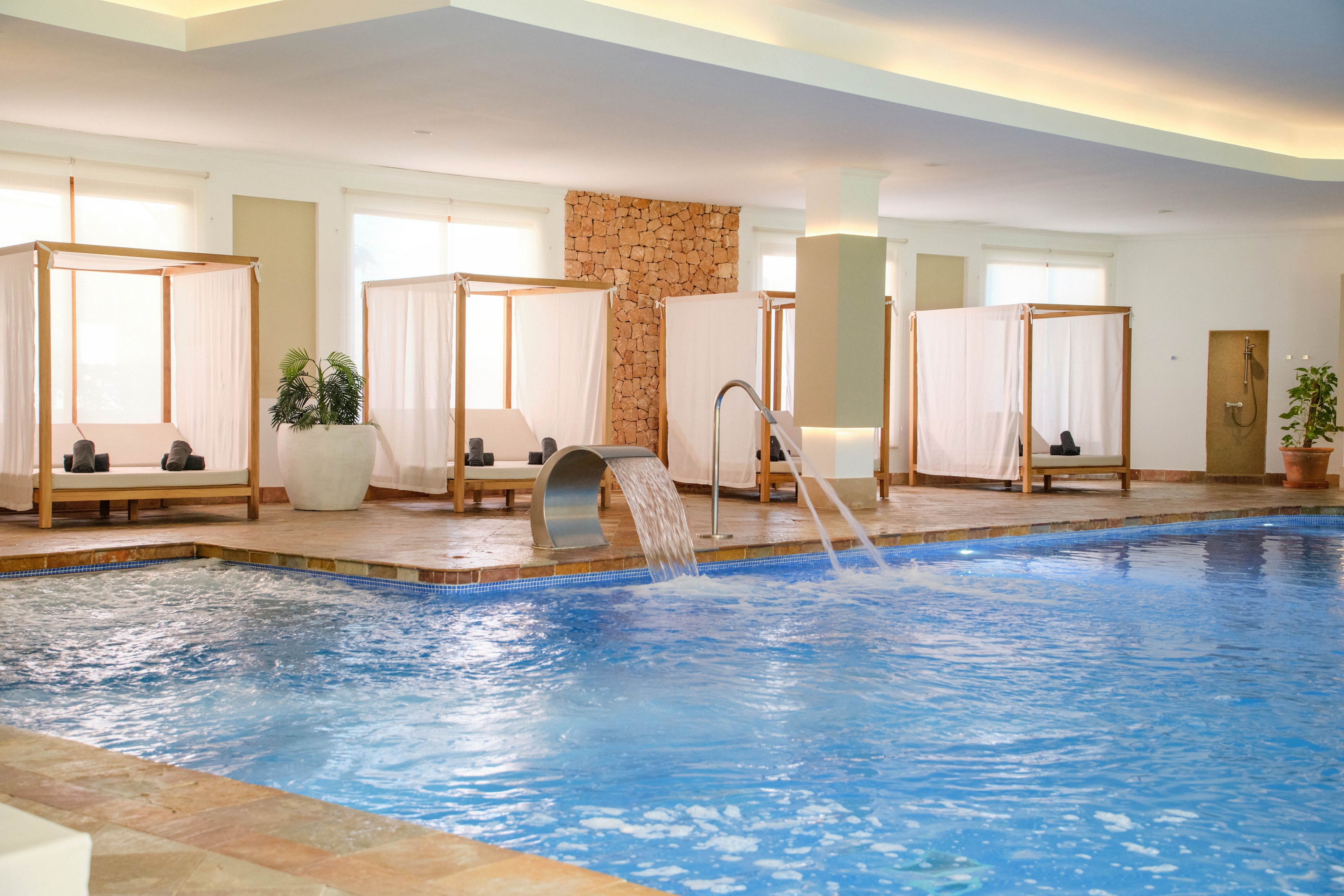 An indoor swimming pool features clear blue water and modern spa-style amenities. Wooden-framed cabanas with white curtains line the far side of the pool, each furnished with dark cushions. Water cascades from a stainless steel fountain and metal handrails lead into the pool. Soft natural light from large windows and warm ceiling lighting create a calm, relaxing atmosphere.