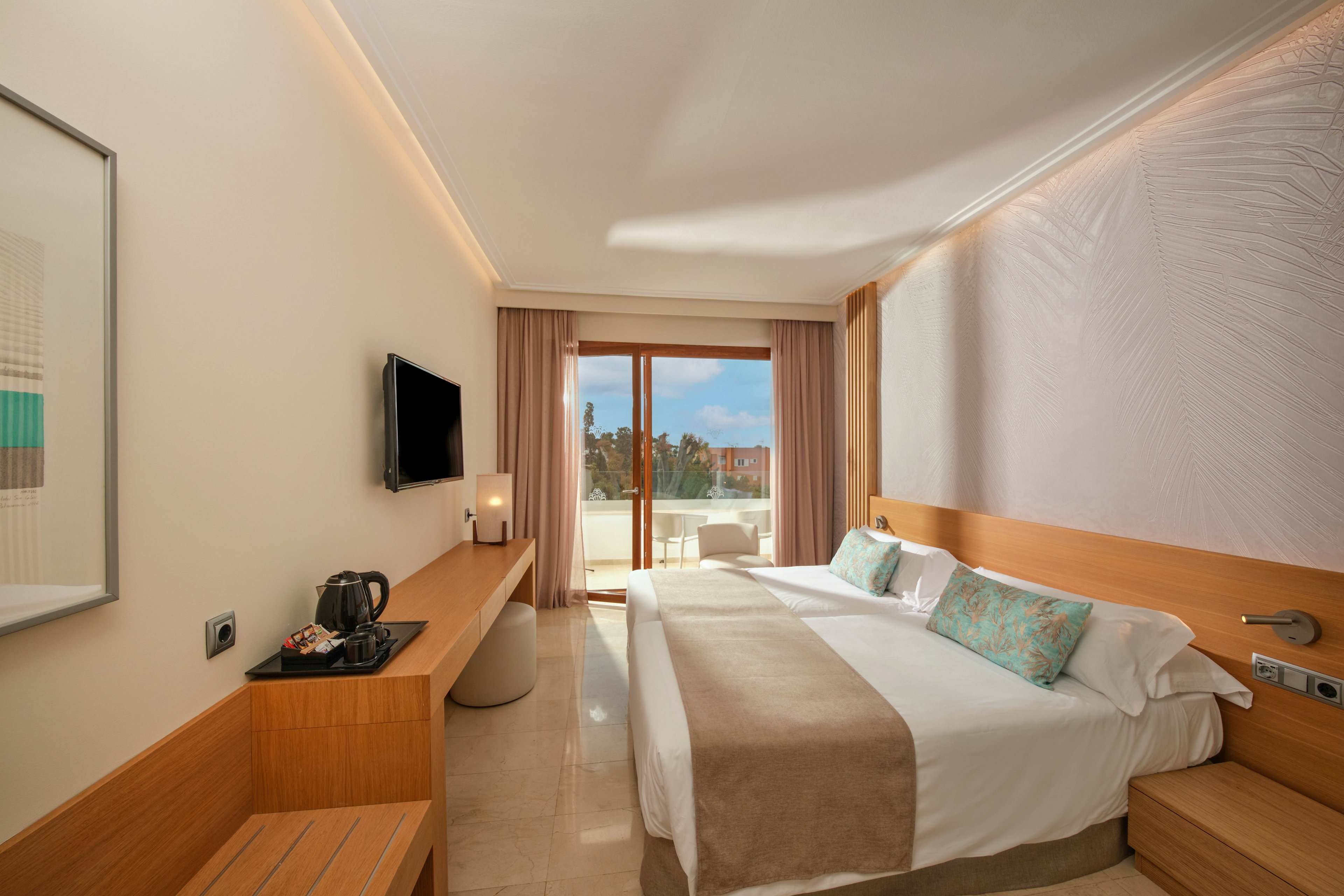 A contemporary hotel room features a large neatly made bed with neutral bedding and decorative pillows. A wall-mounted flat-screen television faces the bed, with a wooden desk holding a kettle and cups. Soft lighting and pale walls create a calm, minimalist atmosphere. Floor-to-ceiling glass doors open onto a balcony with outdoor seating and a view of trees under a blue sky.