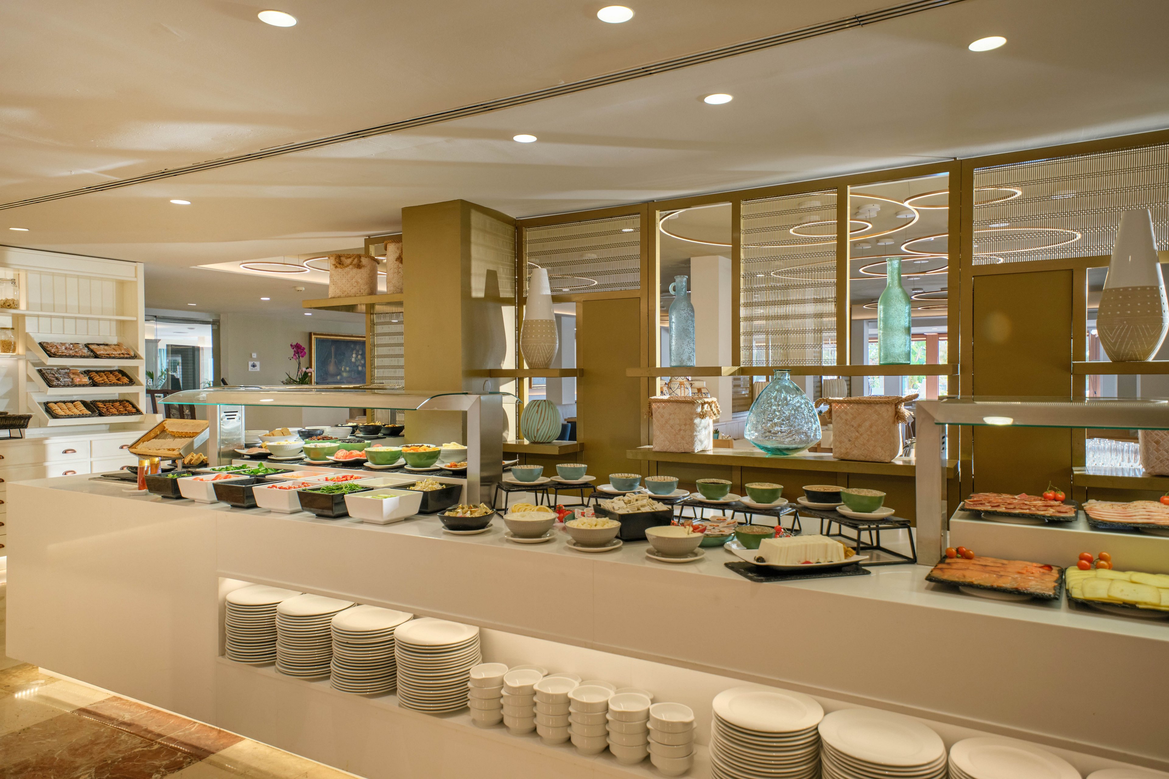 A spacious self-service buffet counter is set up in a modern dining area. Various plates and bowls display salads, cold cuts, and other prepared foods in an orderly arrangement. Stacks of white plates and bowls are neatly organized at the front of the counter for guests. Warm lighting, contemporary decor, and reflective surfaces create an inviting and upscale atmosphere.